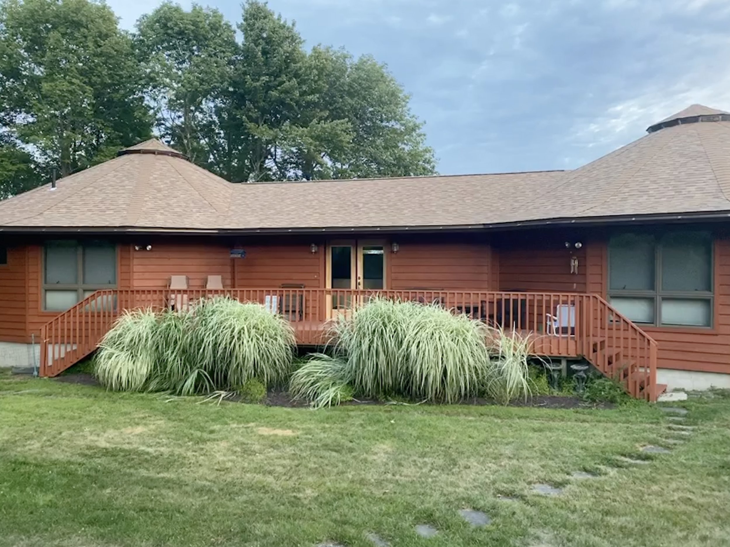 Back of a red wooden house with a deck, steps leading down to a grassy yard, and large ornamental grass plants in front.