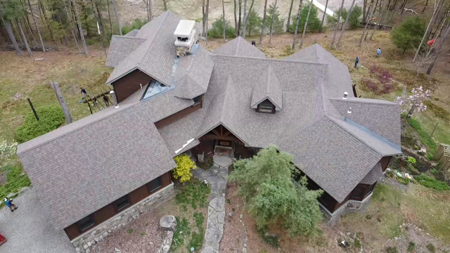 Aerial view of a large house with multiple gray roofs surrounded by trees and a landscaped yard.