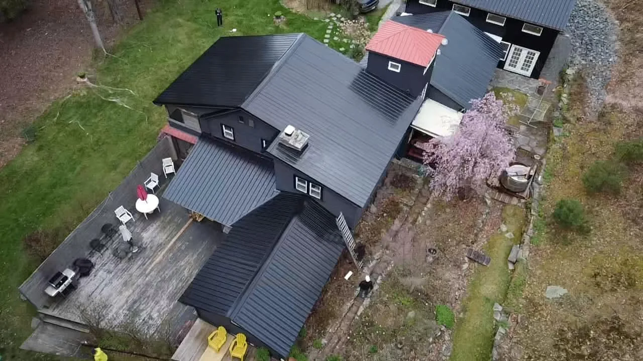Aerial view of a black house with a large deck, outdoor furniture including a table with an umbrella, and a flowering pink tree in the backyard.