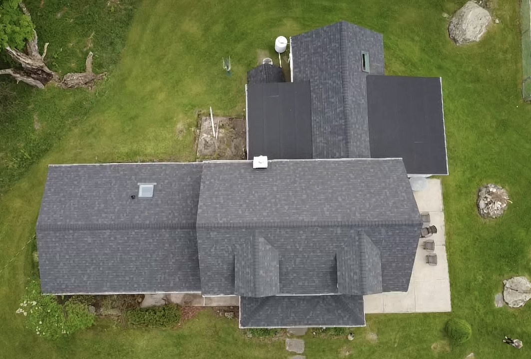 Aerial view of a house with a gray roof, surrounded by a green lawn and some rocks, trees, and outdoor furniture.