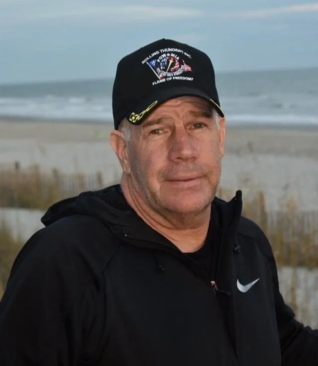 A man in a black Nike jacket and a black cap with an embroidered patch, standing outdoors near the beach with sand dunes and a cloudy sky in the background.