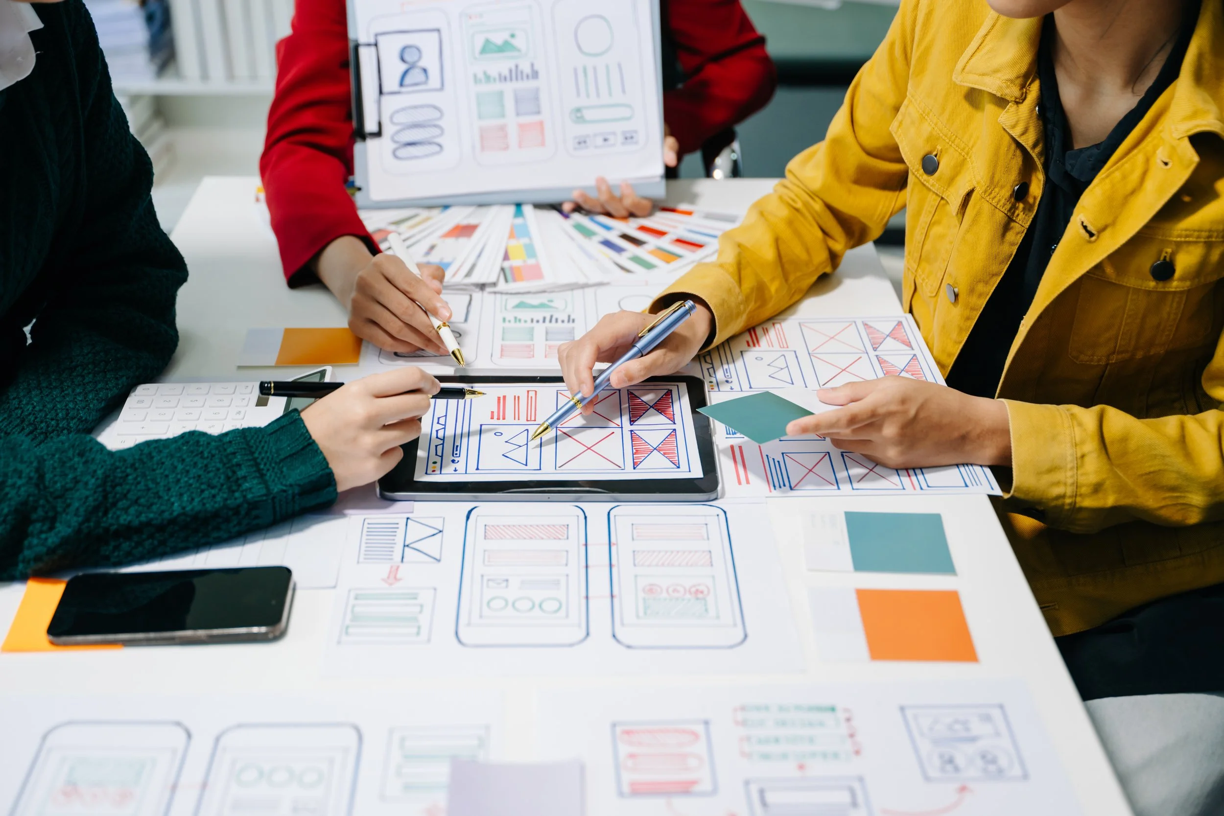 People working on a collaborative project, using a tablet and various design sketches and color samples spread out on the table.