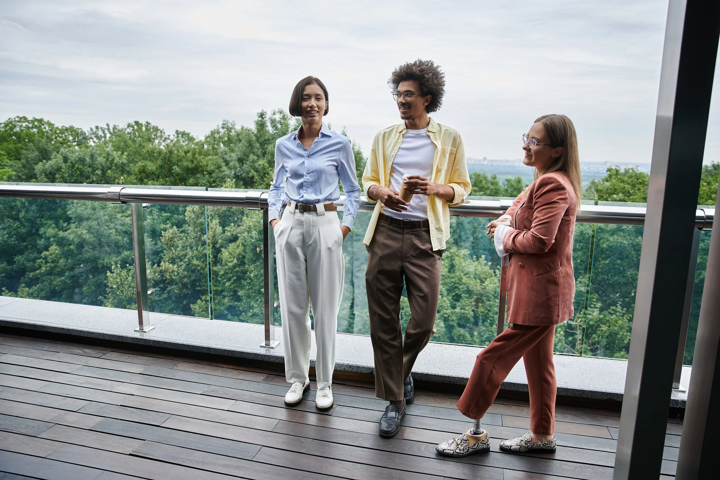 Three people standing on a balcony with a forest view, engaging in conversation and holding coffee cups.