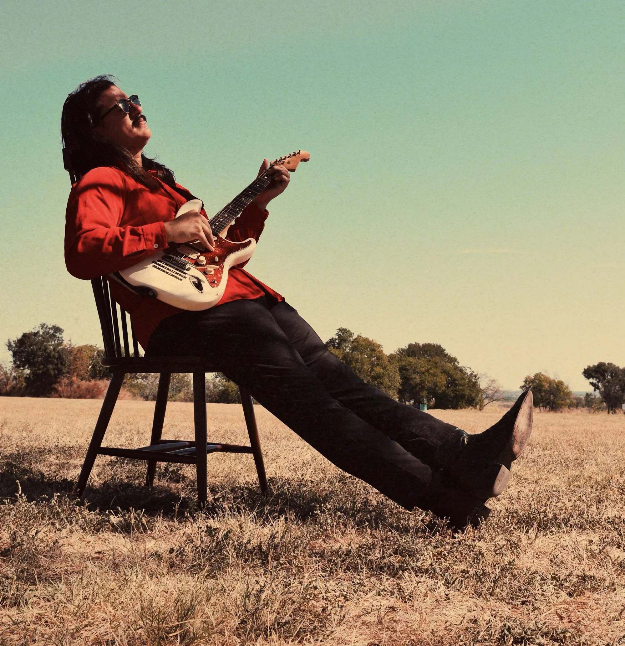 A man with long hair, a mustache and sunglasses sitting on a chair outdoors, playing an electric guitar in a field with trees in the background on a clear day.