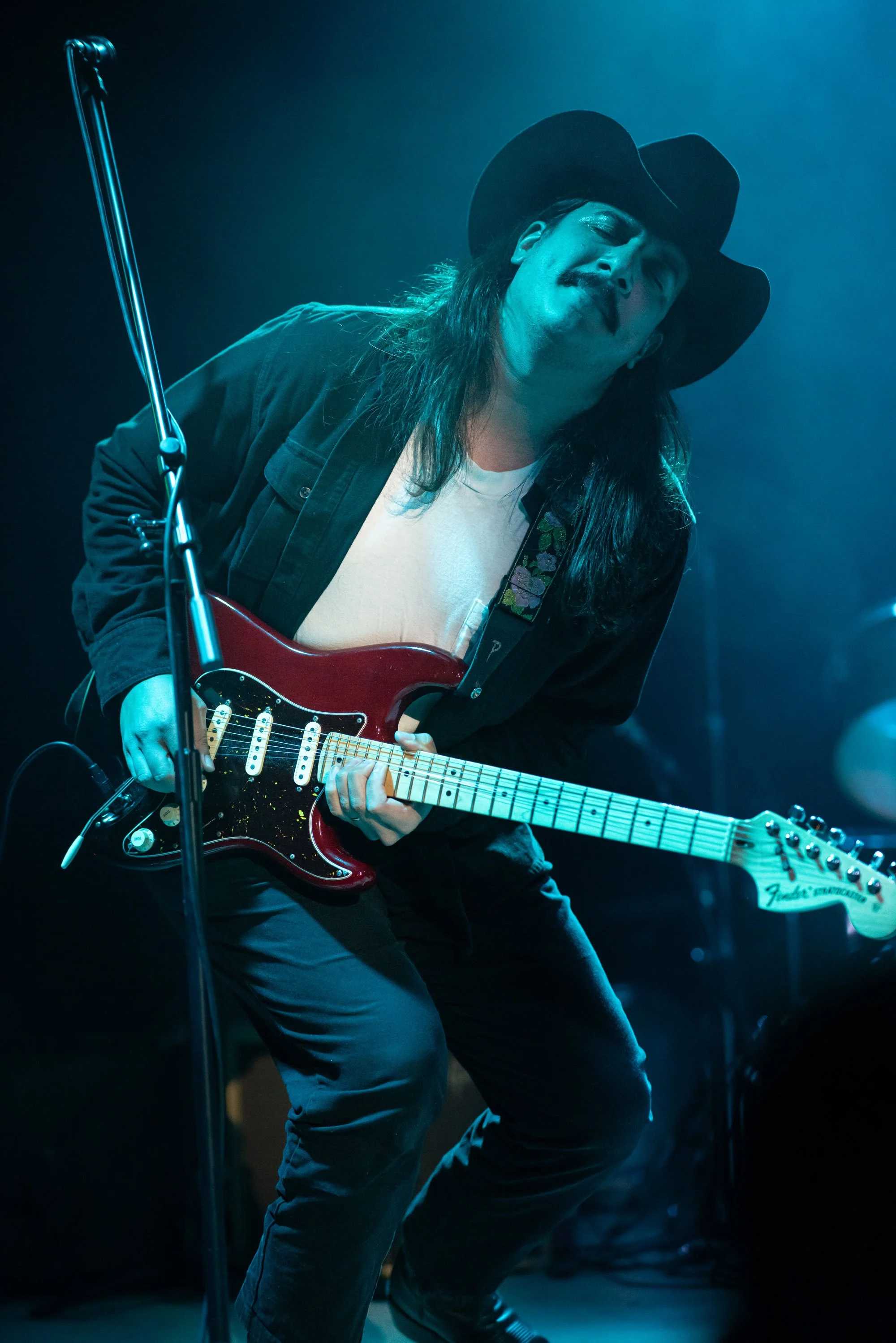 Musician playing electric guitar on stage with a cowboy hat and closed eyes, blue stage lighting, microphone stand in foreground.
