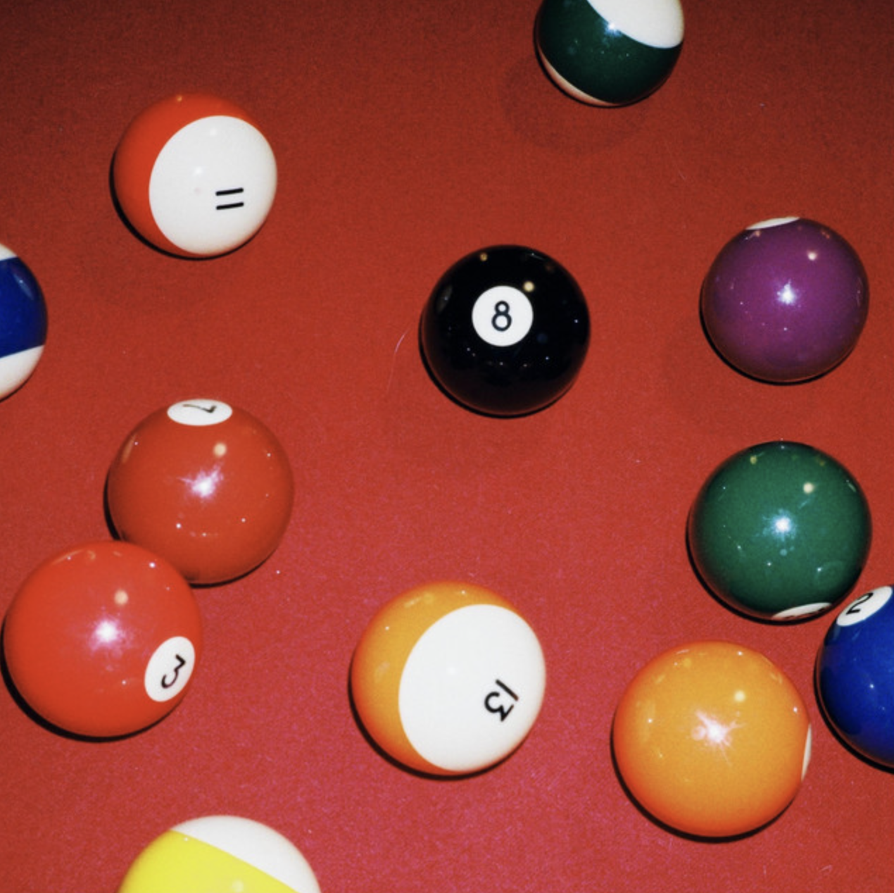 A close-up of various colored pool balls scattered on a red pool table, including the 8 ball, the 3 ball, the 13 ball, and the 7 ball.