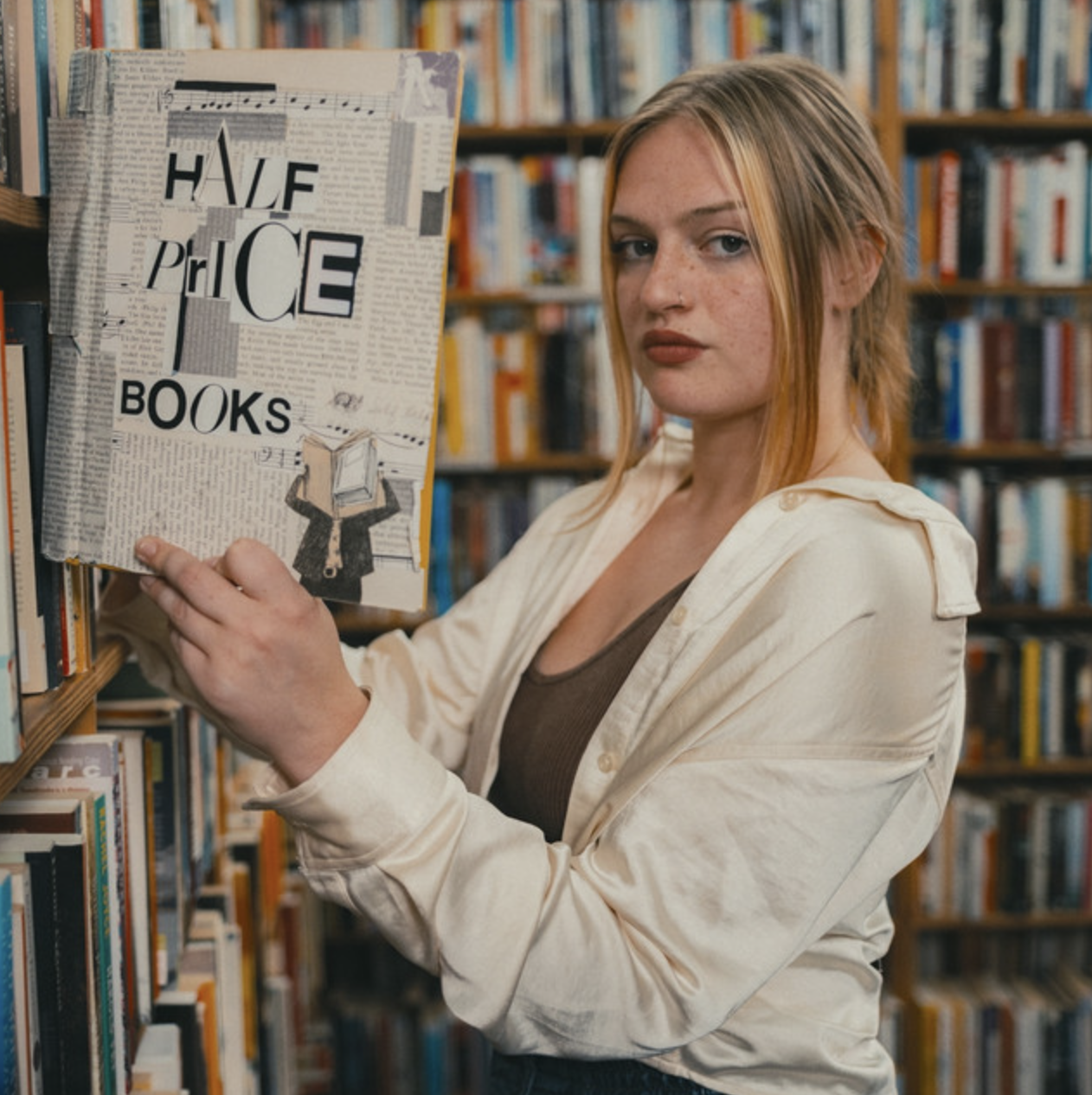 A young woman with blonde hair and freckles holding a book in a library. The cover of the book says "Half Price Books" with a collage-style design, including text and an illustration of a person reading.