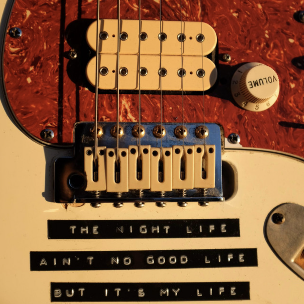 Close-up of an electric guitar with a red tortoiseshell pickguard, white pickups, and a bridge with adjustable saddles. There is a sticker with lyrics that reads, "The night life ain't no good life, but it's my life." A white control knob labeled "VOLUME" is visible on the right side of the image.