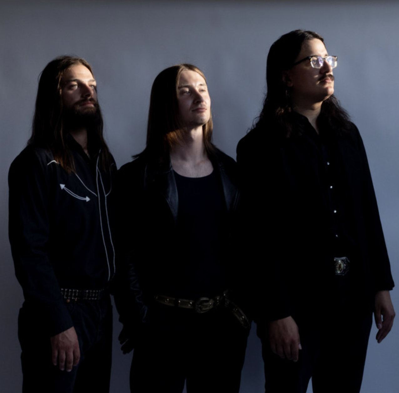 Three men with long hair standing against a plain background, dressed in dark clothing, with serious expressions.