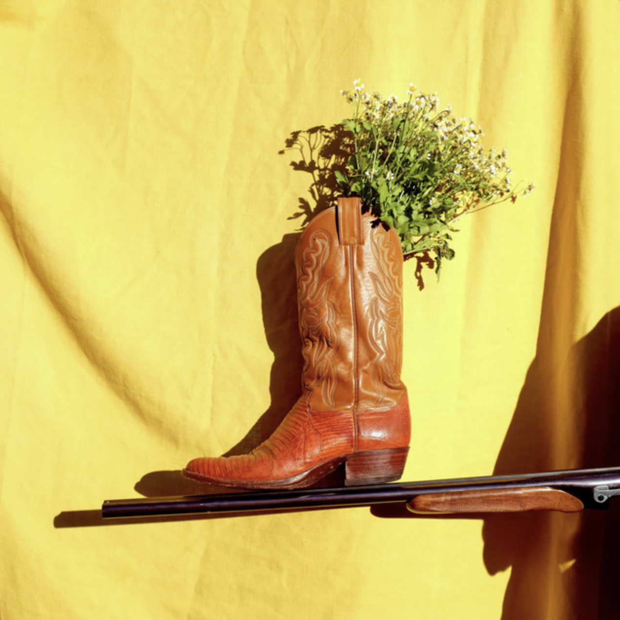 A vintage cowboy boot used as a vase, filled with green foliage and small white flowers, against a yellow background.