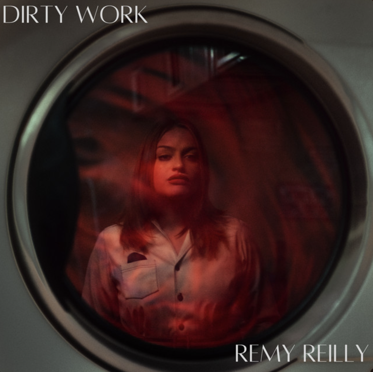 A woman with shoulder-length brown hair looking through a red-tinged washing machine door, with the title "DIRTY WORK" and the name "REMY REILLY" on the image.