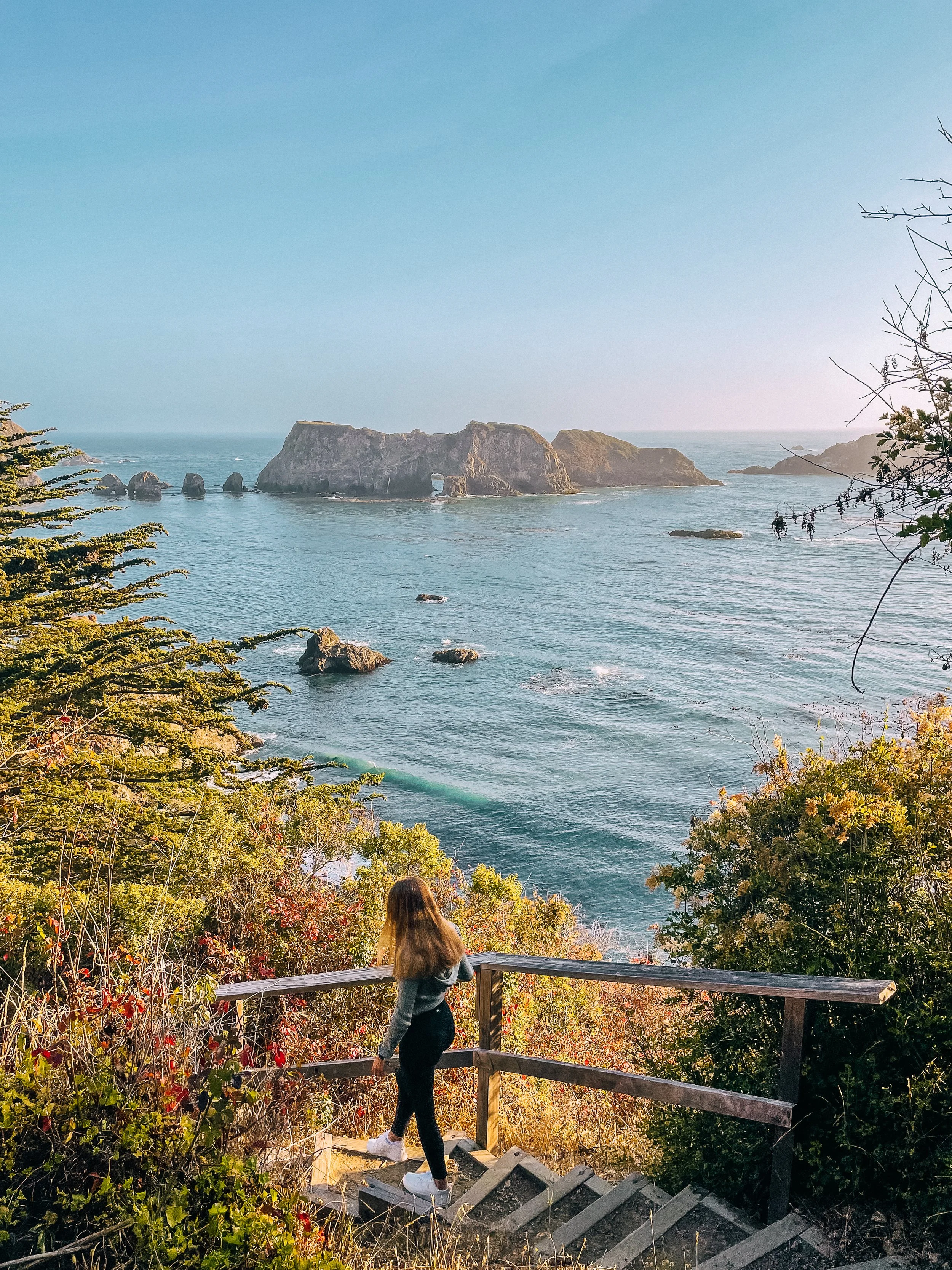 A woman with long hair looking out over the ocean from a wooden staircase, with rocky islands in the distance and lush greenery surrounding the area.