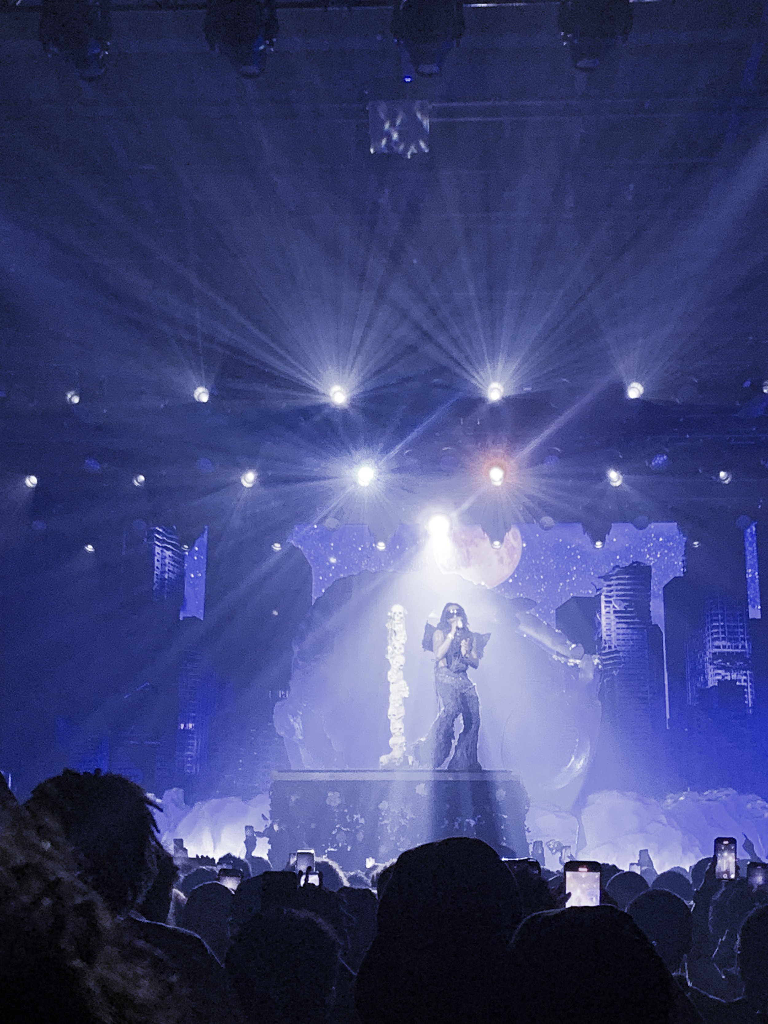 Singer performing on stage with dramatic lighting and celestial background, attended by a crowd with illuminated phones.