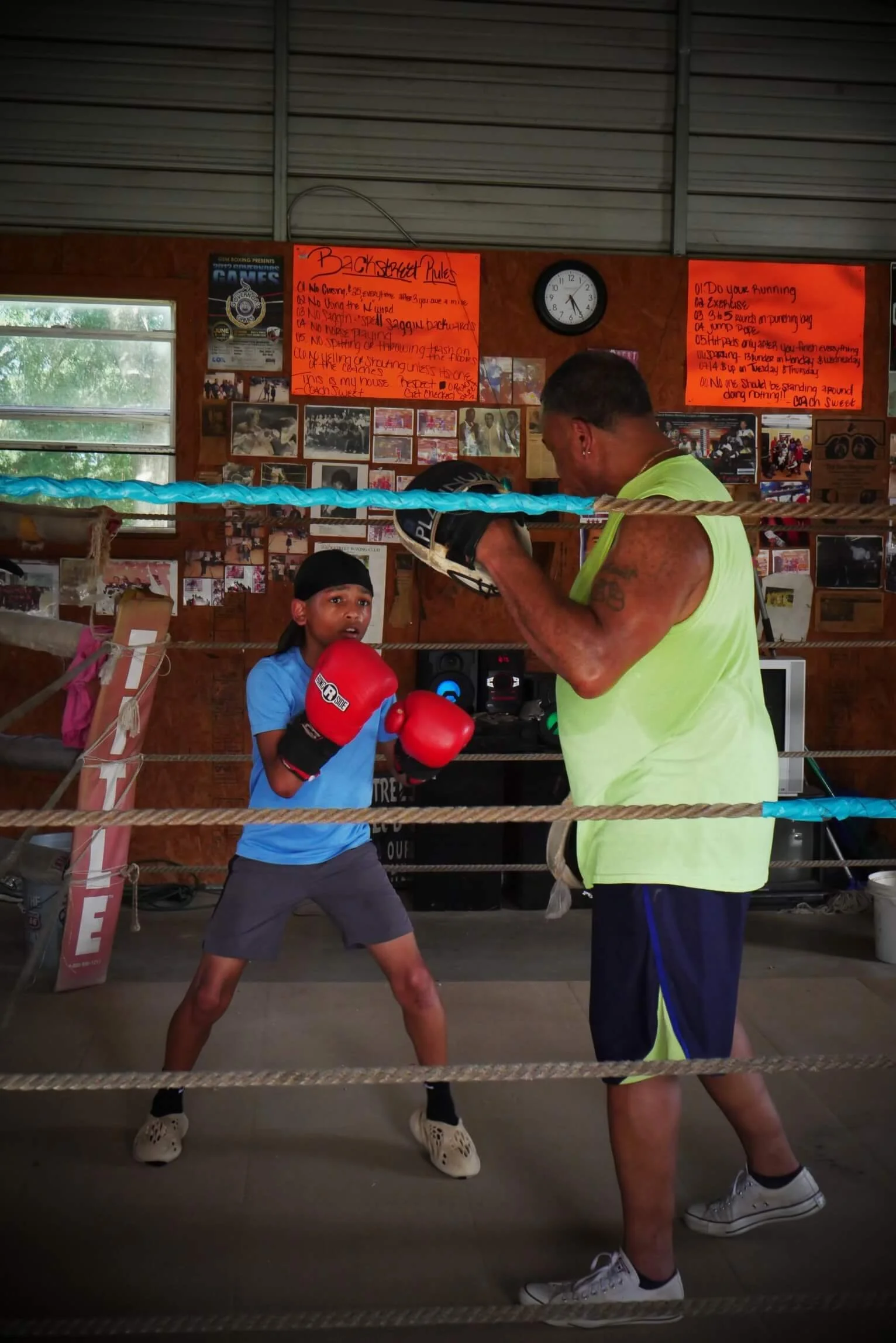 A young girl sparring with a coach in a boxing ring, wearing red boxing gloves and a blue shirt, while the coach practices boxing techniques holding a protective mitt. The background features photo displays, orange signs with text, and a clock on the wall.