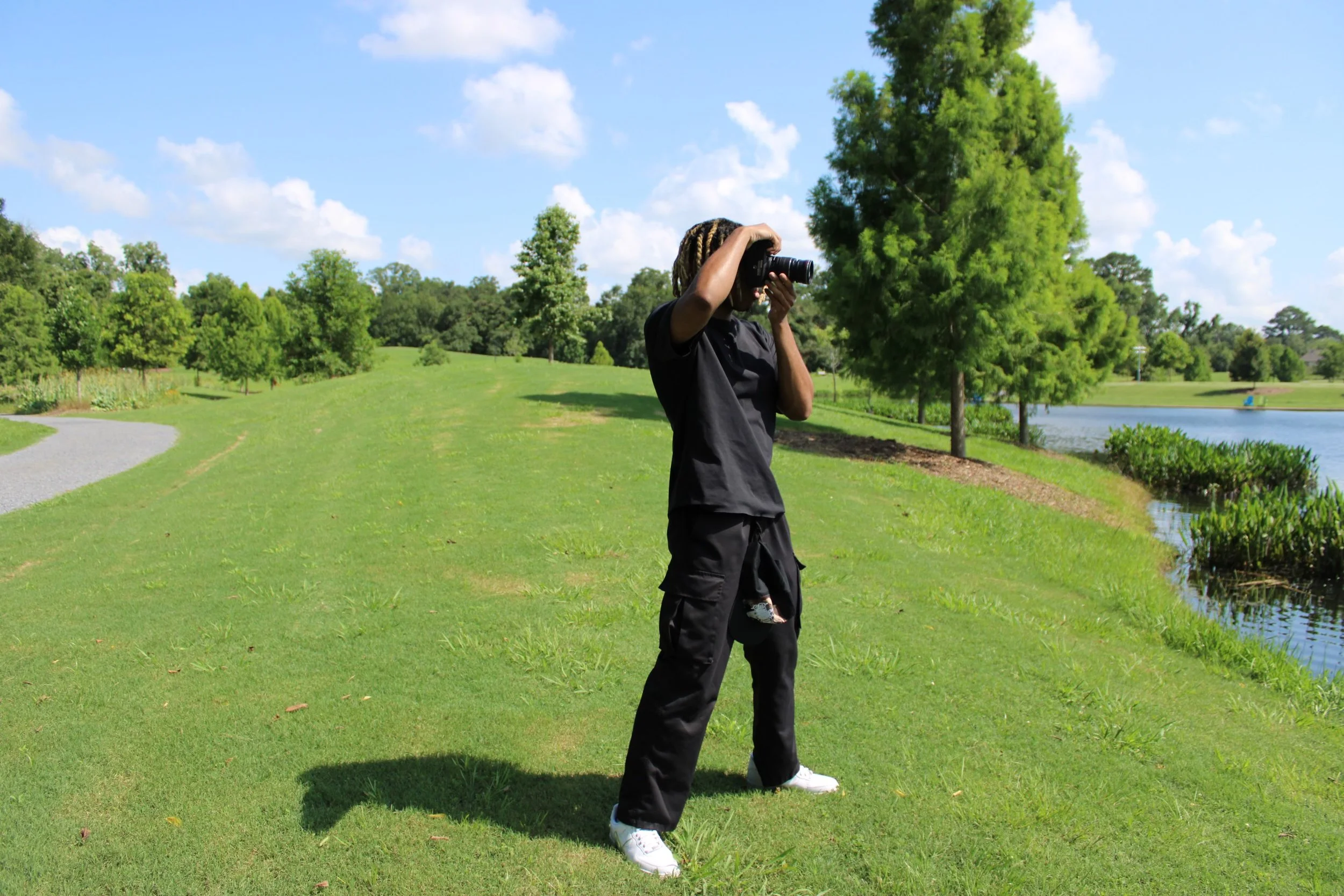 Person taking a photo in a park, standing on grass near a pond with trees and blue sky in the background.