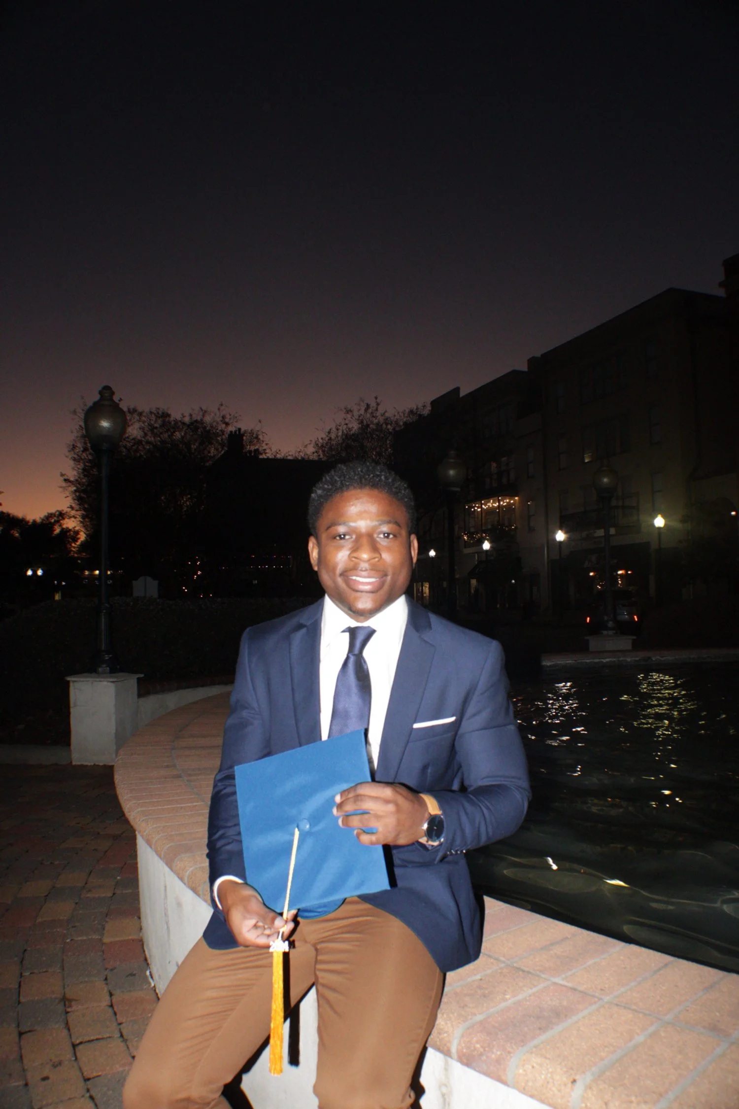 Person in formal attire holding a graduation cap, sitting by a fountain with a cityscape backdrop at dusk.