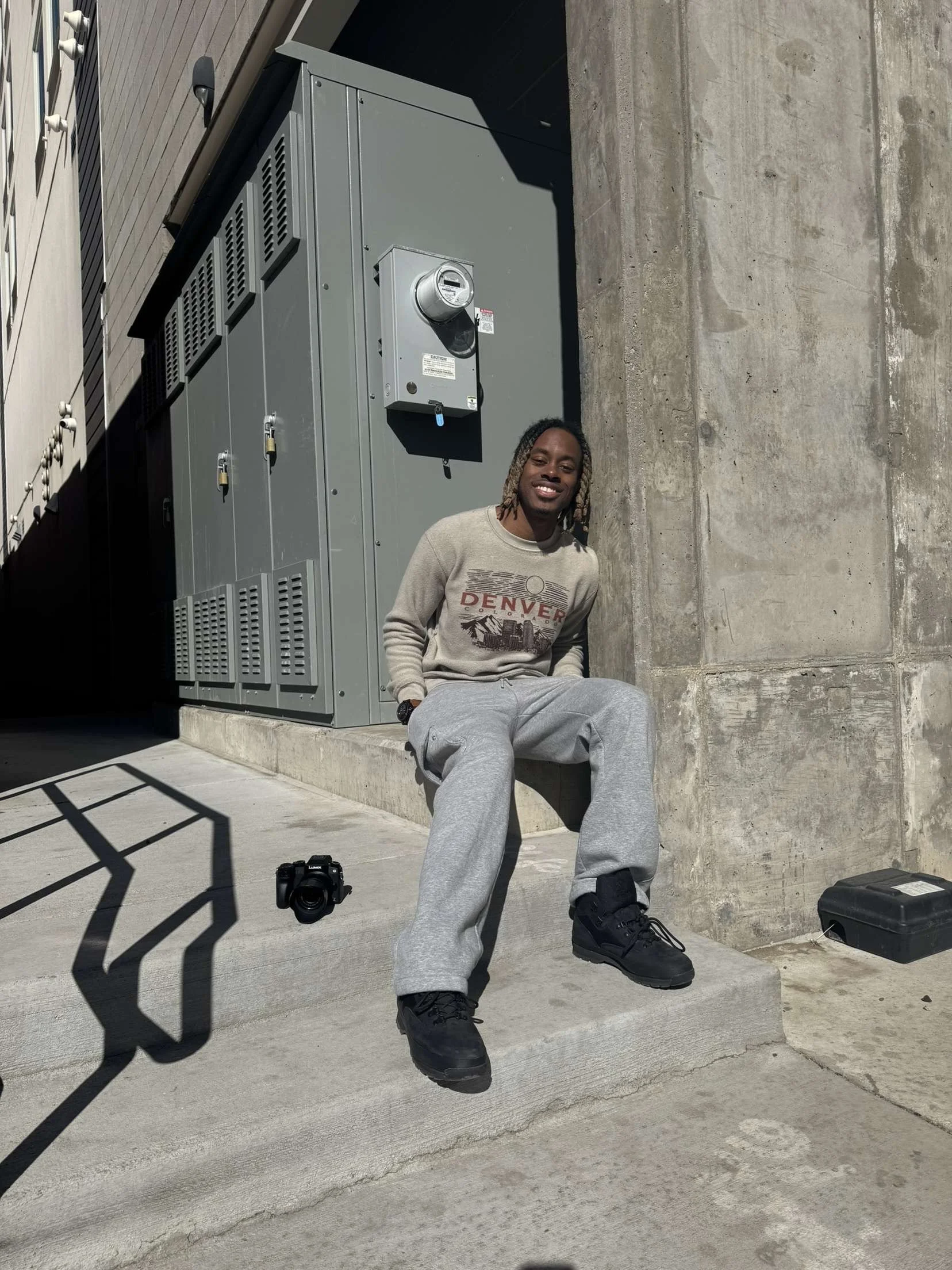 Person sitting on concrete steps next to a camera and a building with an electrical meter, wearing a beige Denver sweatshirt and gray sweatpants.