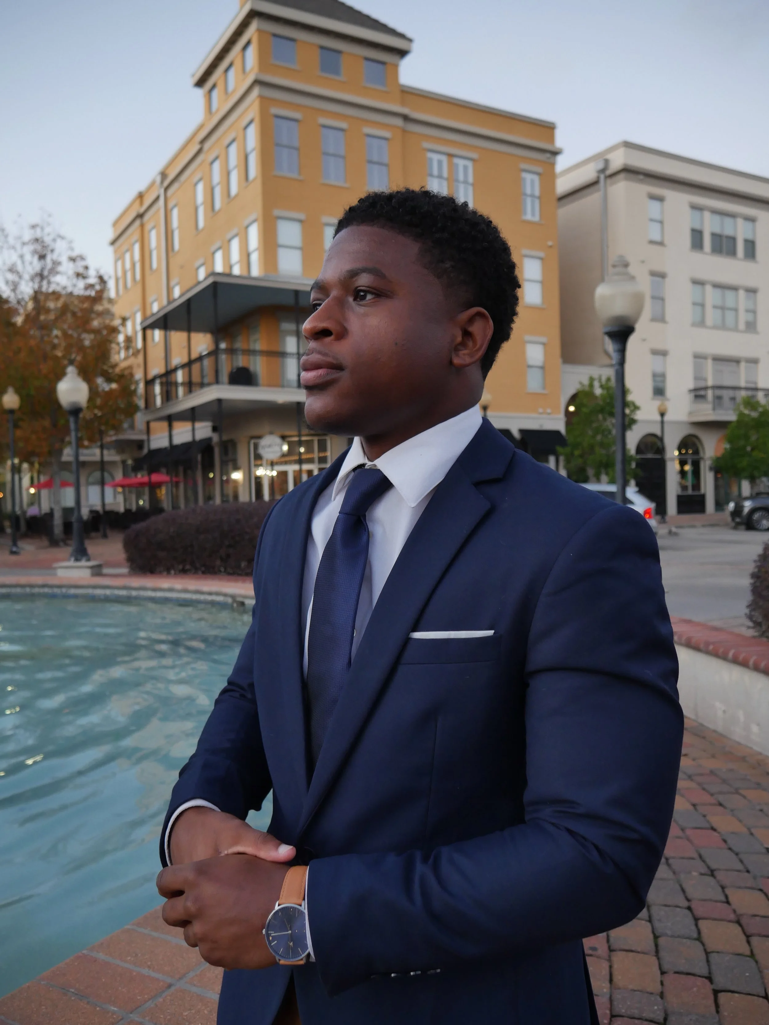 Man in a blue suit standing near a fountain with buildings in the background.