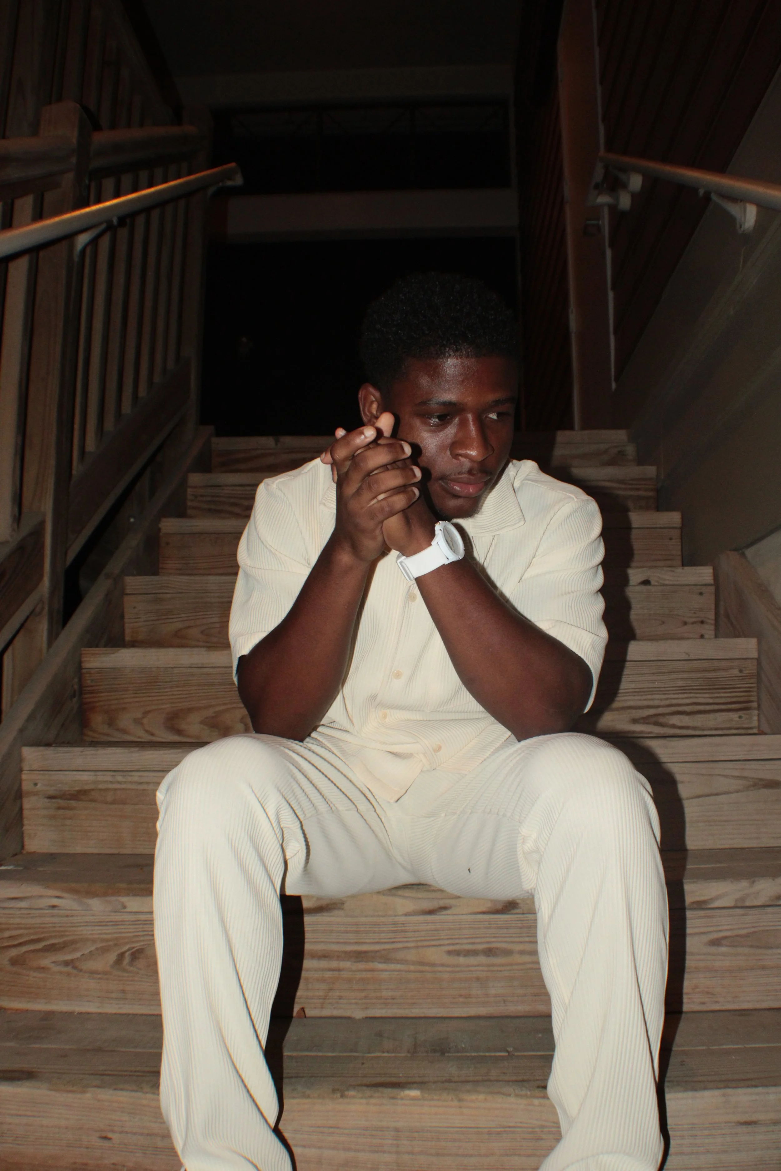 A man sitting on wooden stairs, looking thoughtful and wearing a white outfit and watch.