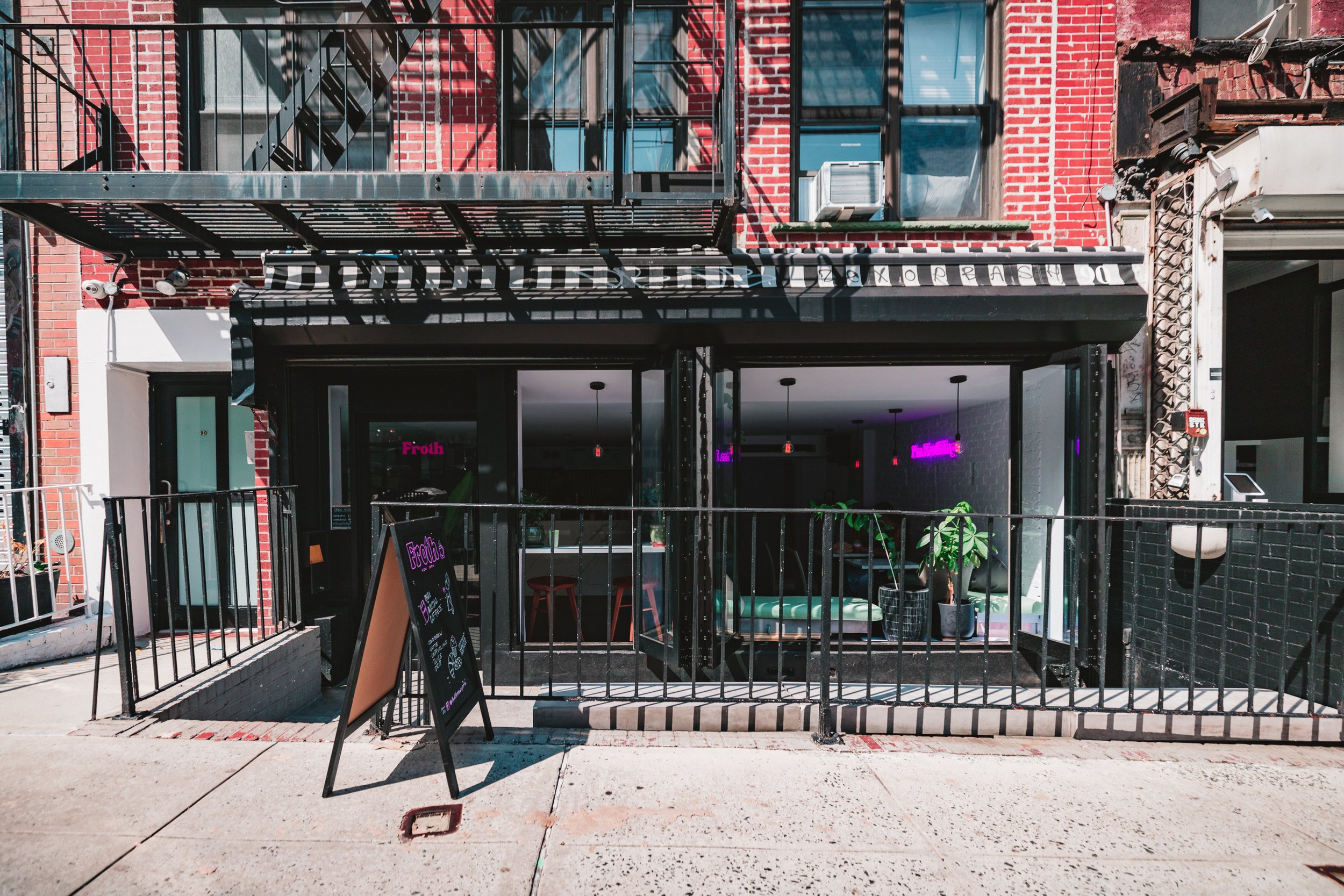 Exterior view of a modern cafe with a glass facade, black framed entrance, neon pink signs, and outdoor seating with green plants in pots.