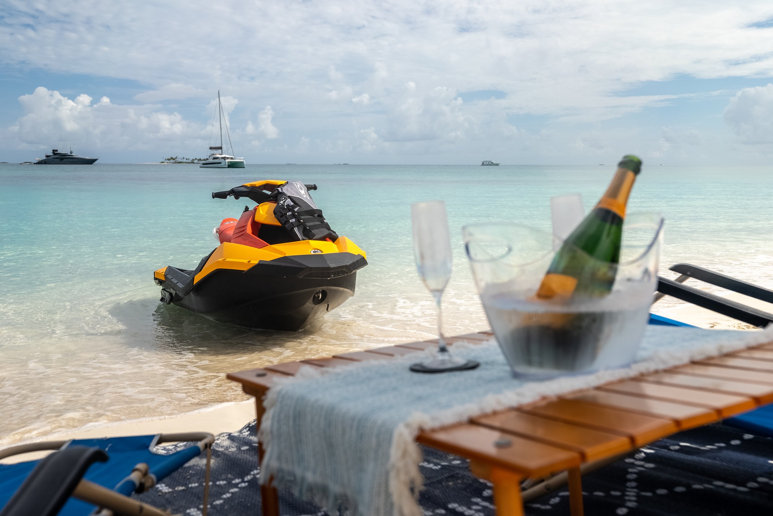 Beach scene with jet ski in shallow water, champagne bottle in ice bucket on a table, and two empty glasses on a sunny day