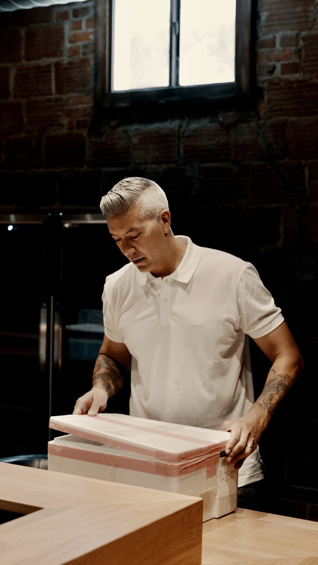 A man with styled hair and tattoos on his arms is opening a cardboard box on a wooden table inside a room with brick walls and a window.