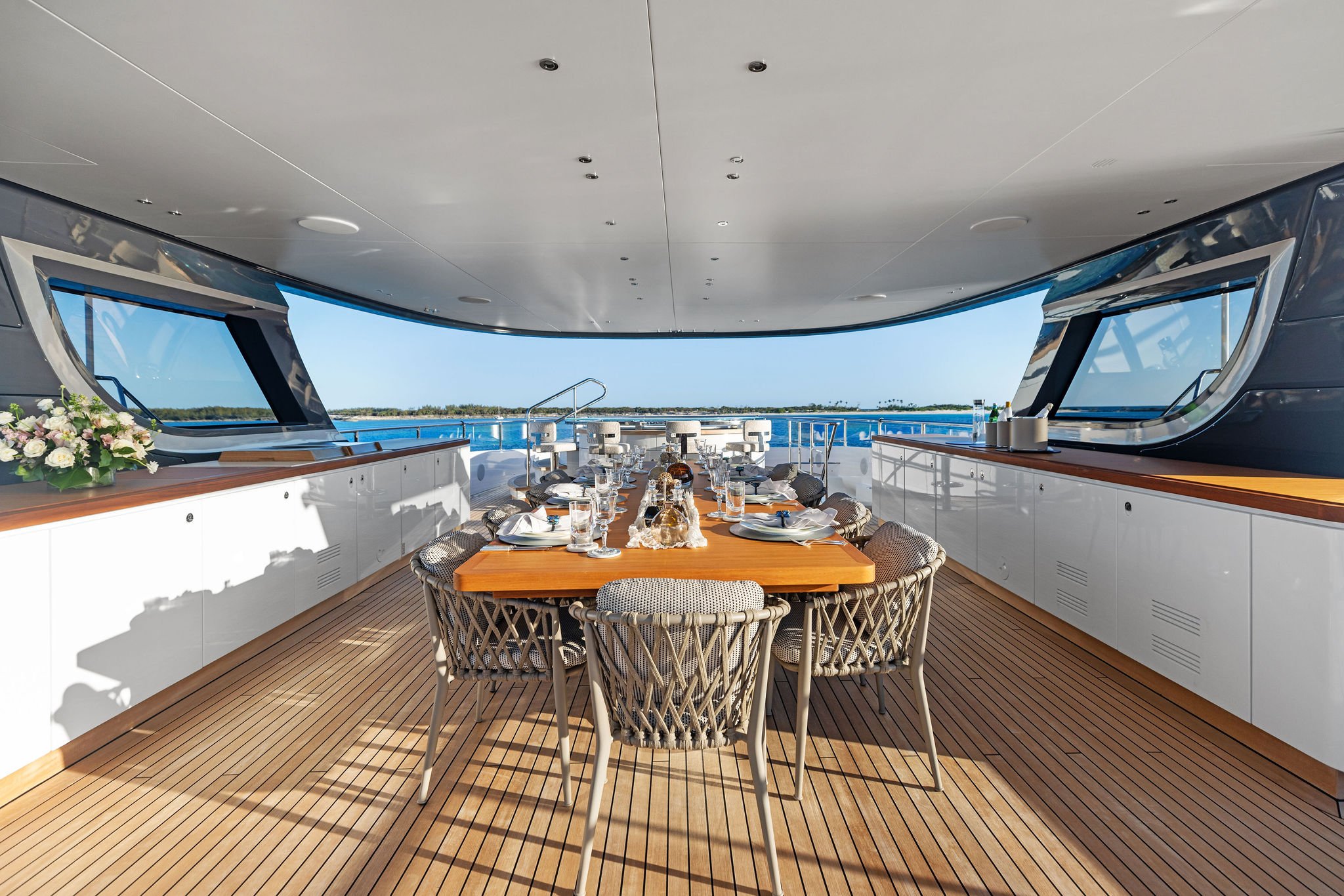 A dining area on a yacht with a wooden table set for a meal, surrounded by wicker chairs, overlooking the ocean with clear skies.