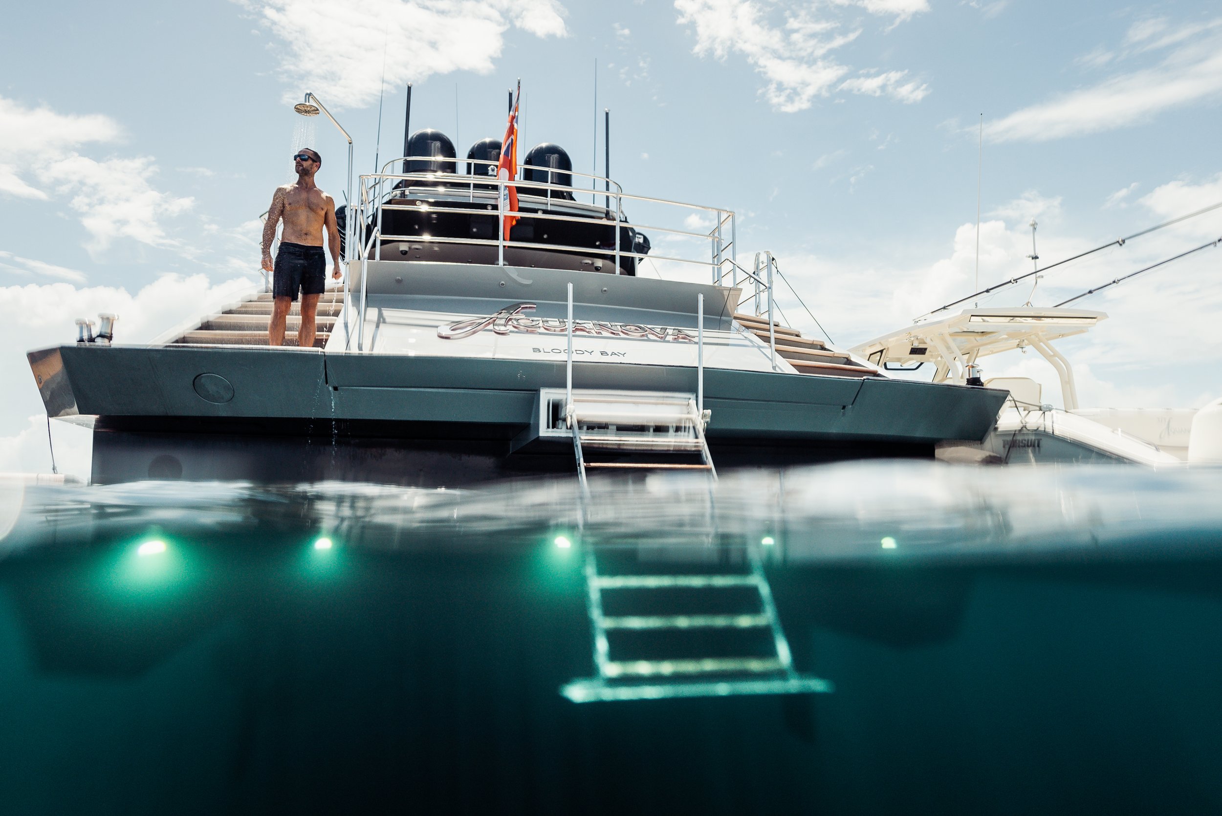 Man standing on the stern of a yacht named Bloody Bay, with steps leading into the water, during daytime with partly cloudy sky.