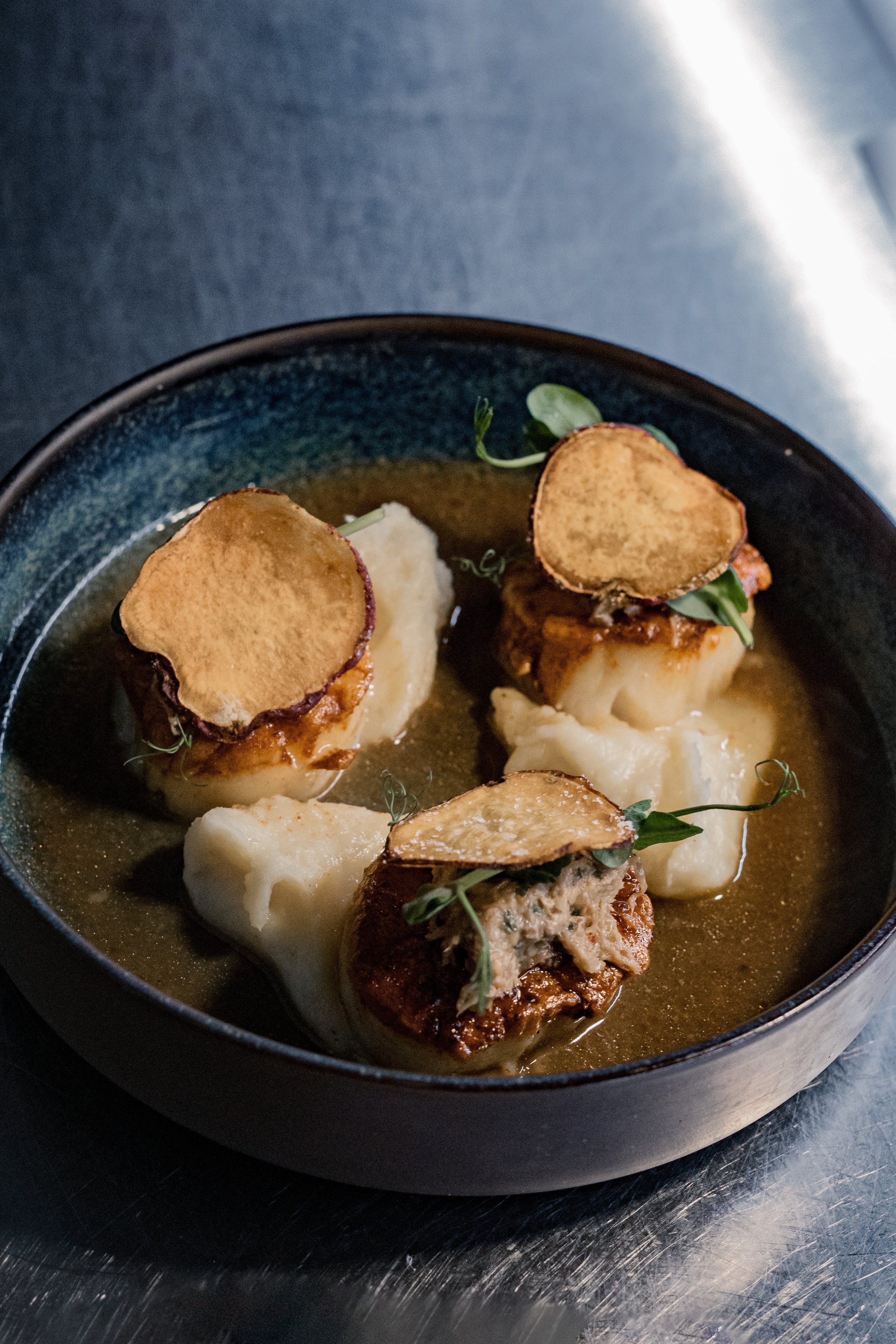 Dish with three meatballs topped with crispy potato chips, served with mashed potatoes and green garnish in a black bowl.