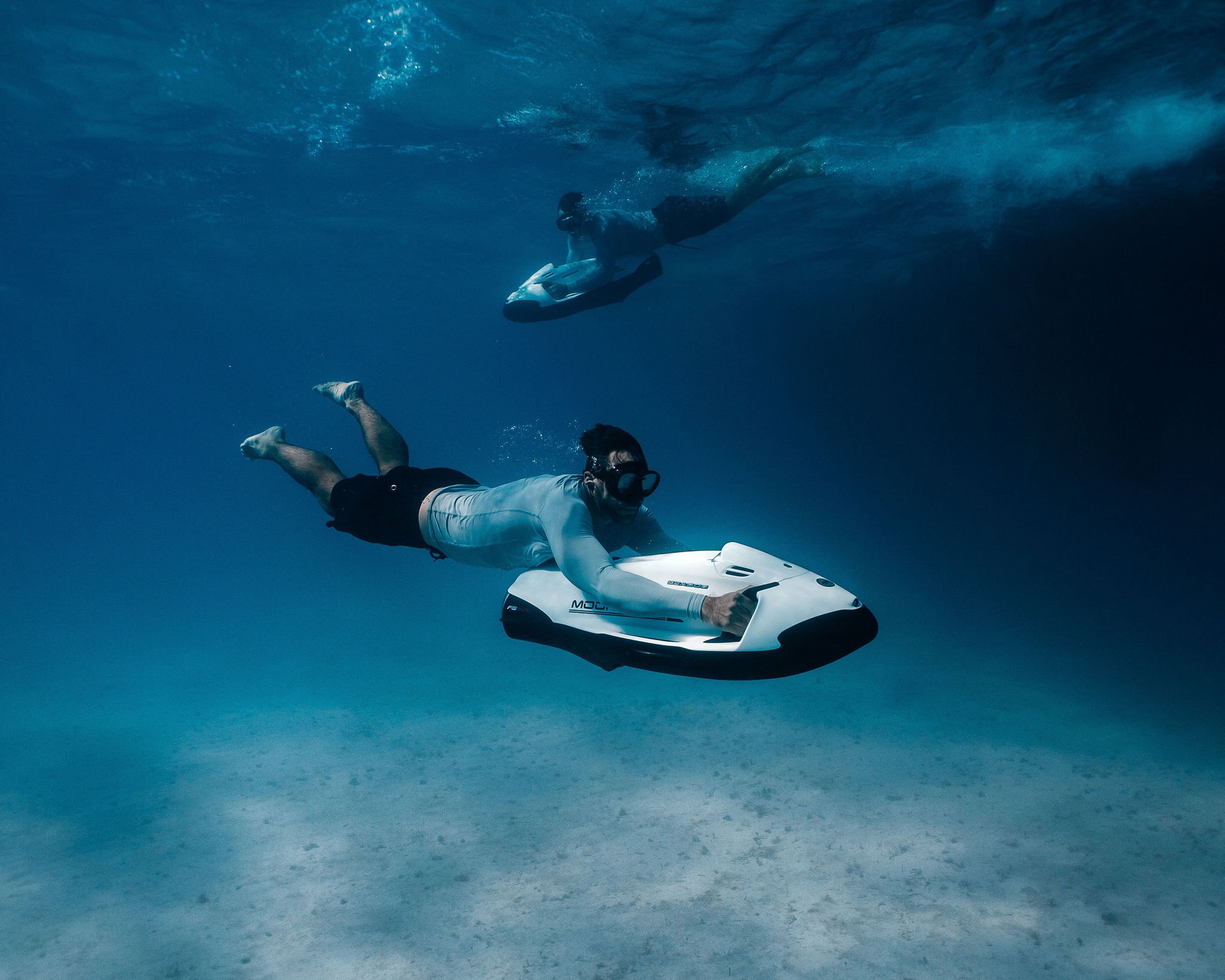 Man swimming underwater with a scooter, wearing goggles, with a person on a paddleboard visible in the background.