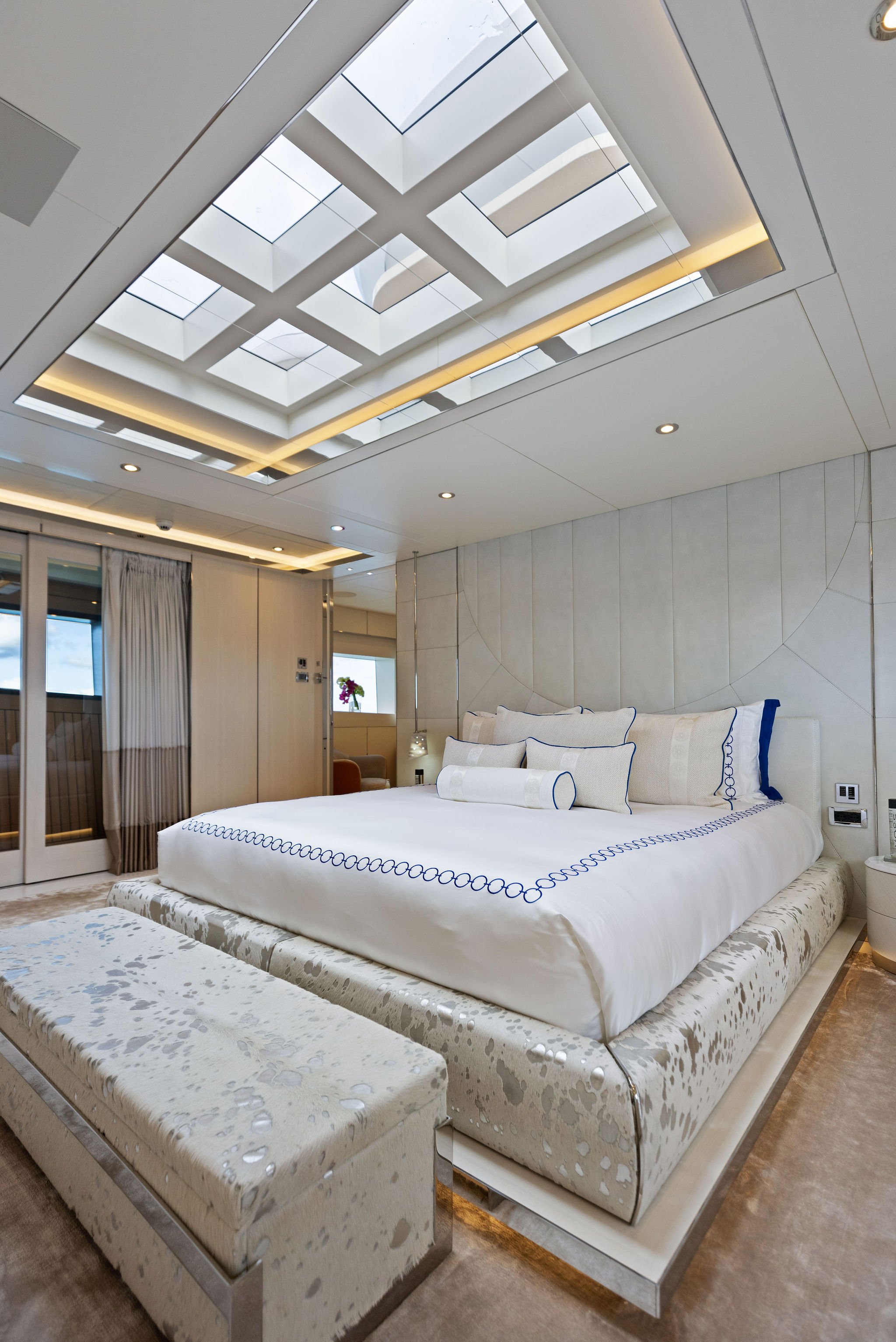 Luxurious hotel bedroom with a large white bed, upholstered bench at the foot, skylight with geometric pattern on the ceiling, and modern decor.