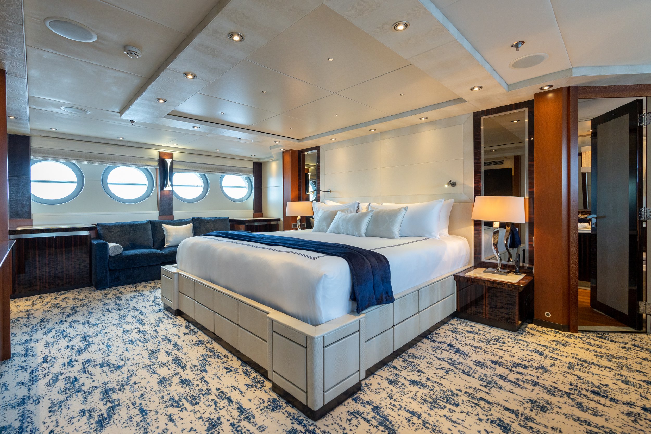 Luxurious modern bedroom with a large bed, a navy blue throw, multiple pillows, bedside lamps, a navy sofa, and circular windows along the wall.