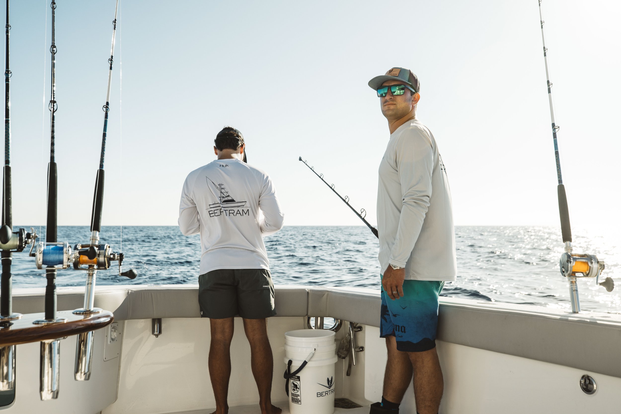 Two men fishing on a boat in the ocean during sunset, with fishing rods and a white bucket.