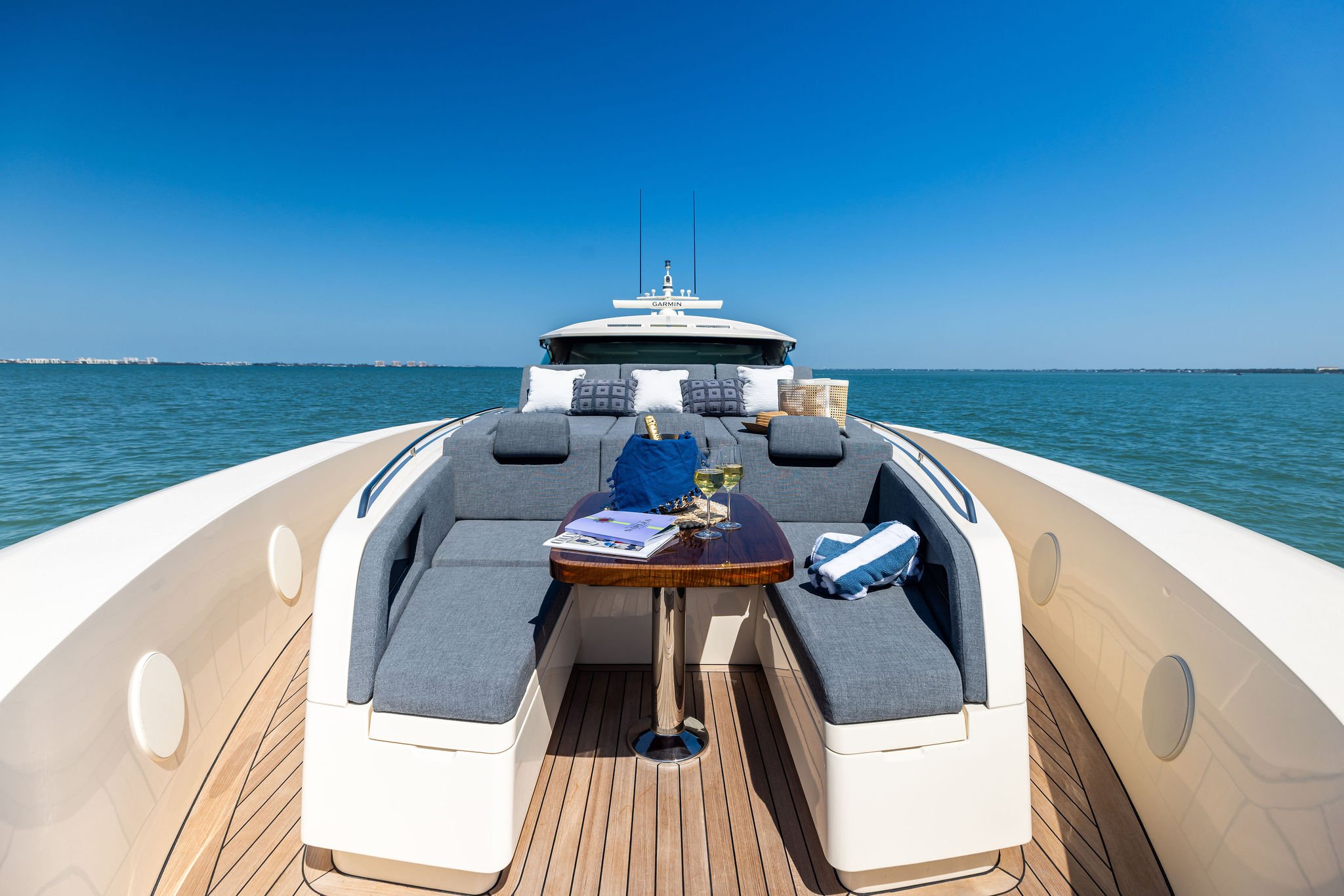 Luxury yacht with gray cushioned seating area on deck, set against a backdrop of blue ocean and sky, with distant shoreline on the horizon.