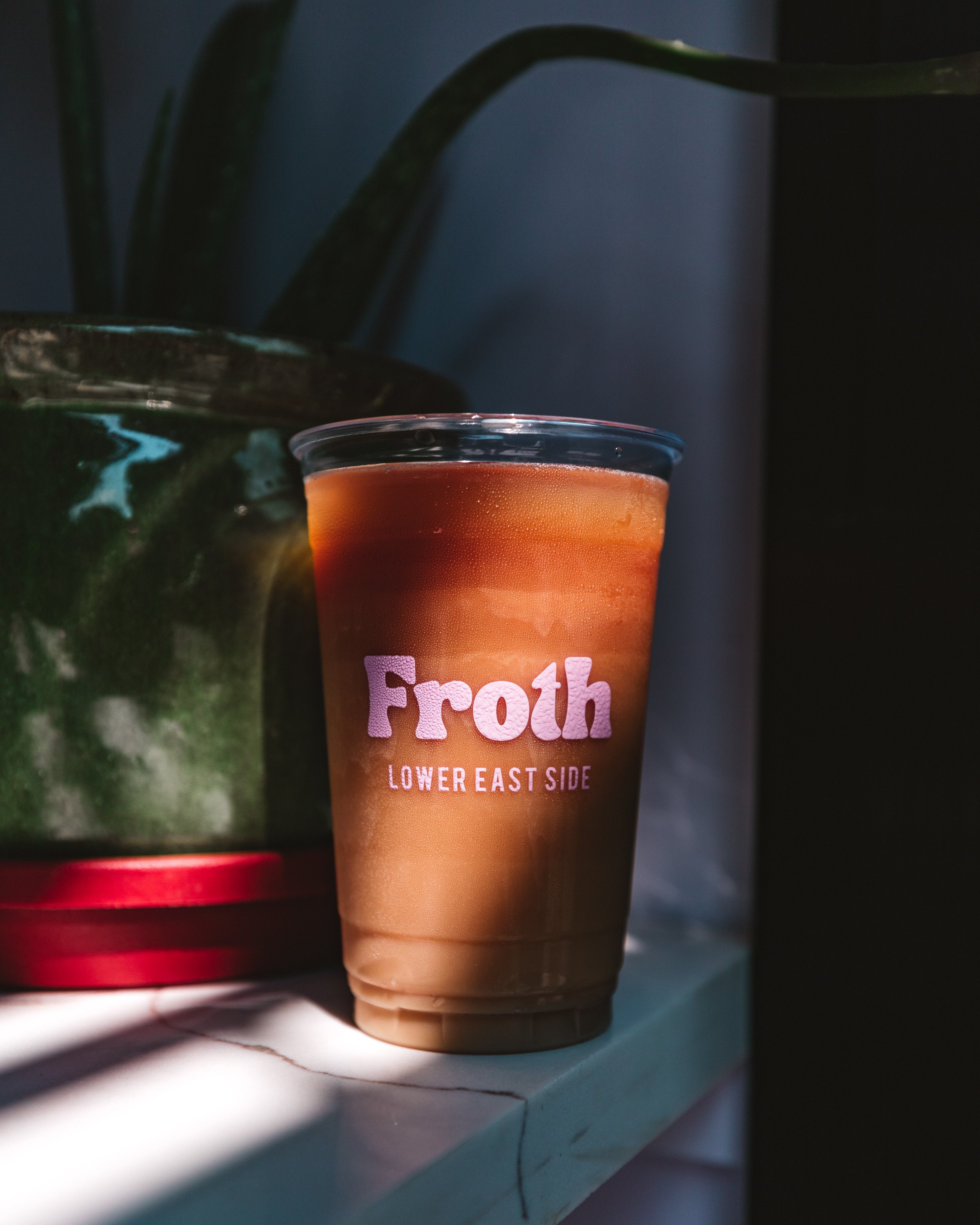 A plastic cup with iced coffee placed on a white marble surface, with the brand name 'Froth' and 'Lower East Side' printed on it, next to a large green plant.