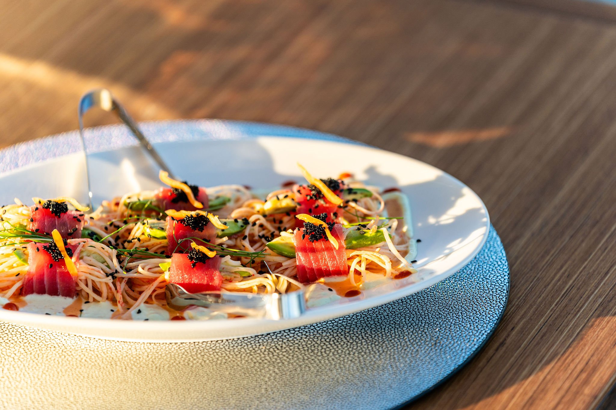 Plate of Asian noodle salad with slices of raw tuna, garnished with green herbs, black sesame seeds, and bright orange zest, placed on a textured silver placemat on a wooden table.