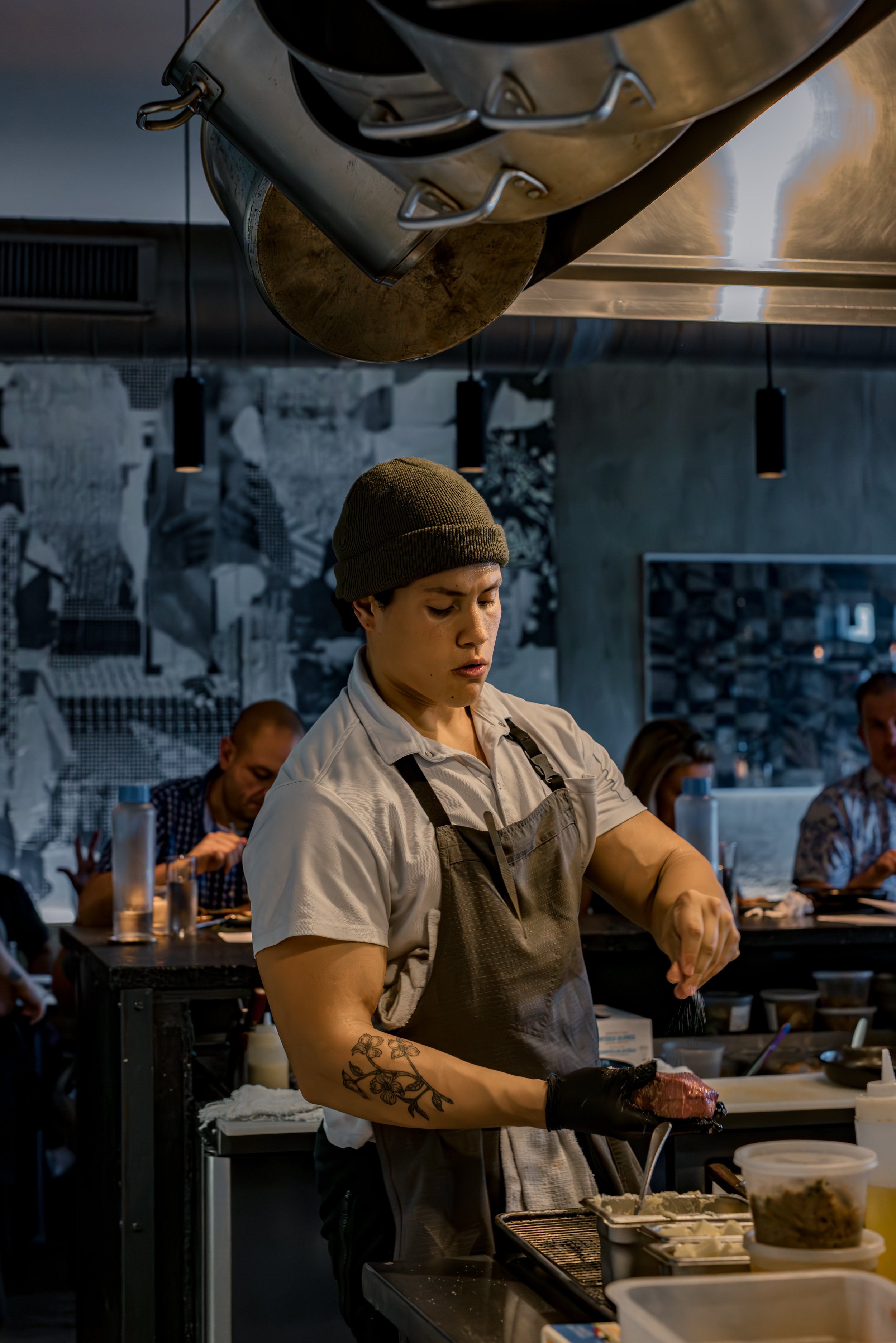 A chef with a tattoo on her arm wearing a brown beanie and a white shirt, preparing food in a modern restaurant kitchen with other staff and customers in the background.