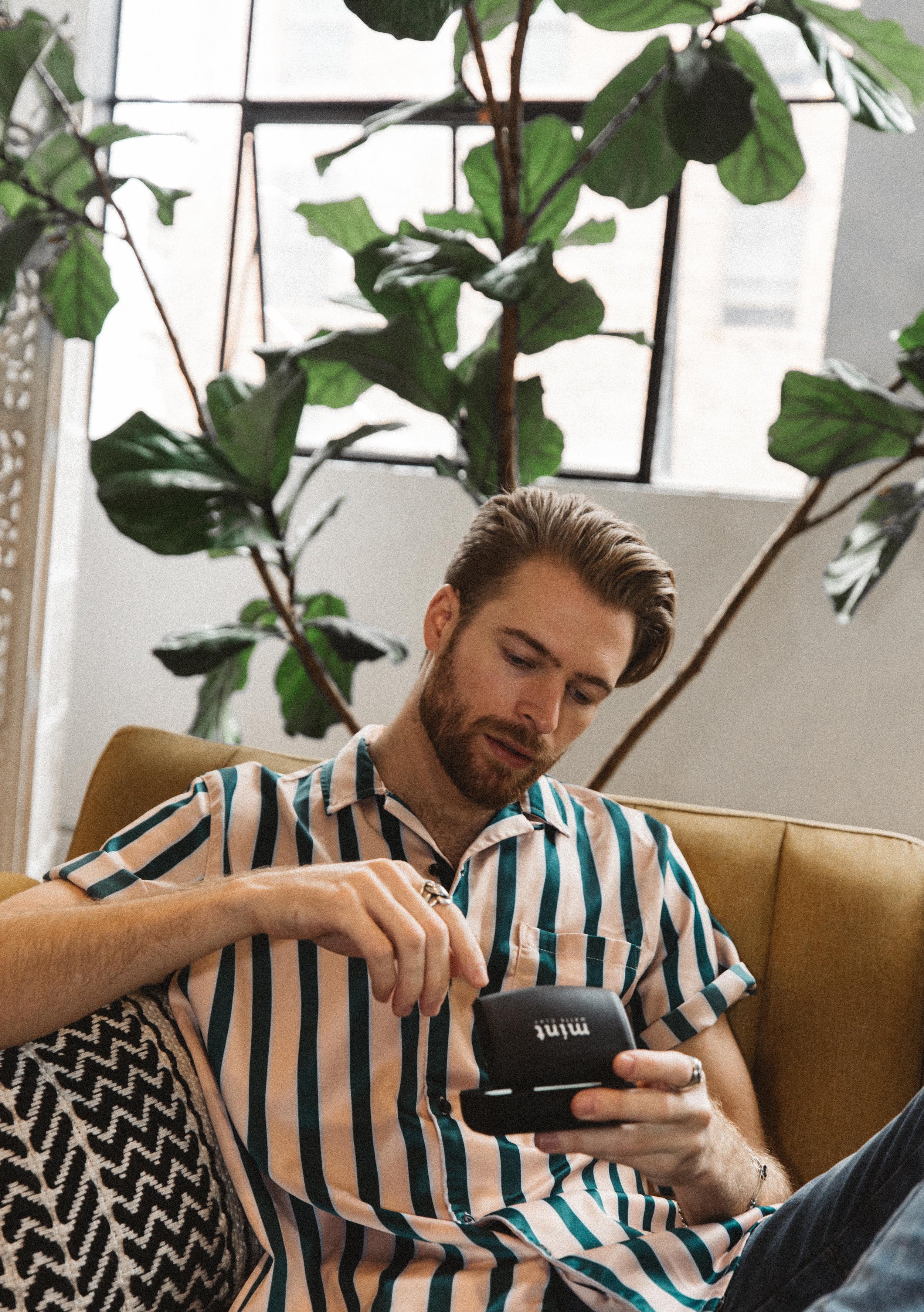A man with a beard and styled hair sitting on a mustard-colored couch, looking at a black portable speaker or radio, with large leafy green plants behind him and a window in the background.