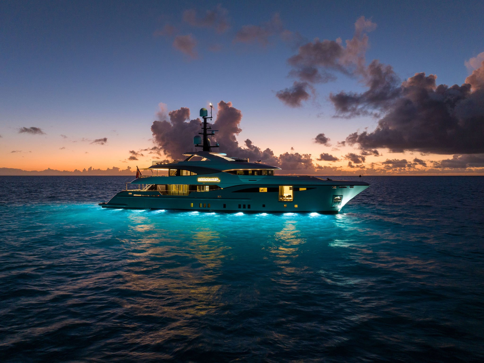 A luxury yacht on the ocean at sunset, illuminated with underwater lights, with a cloudy sky in the background.