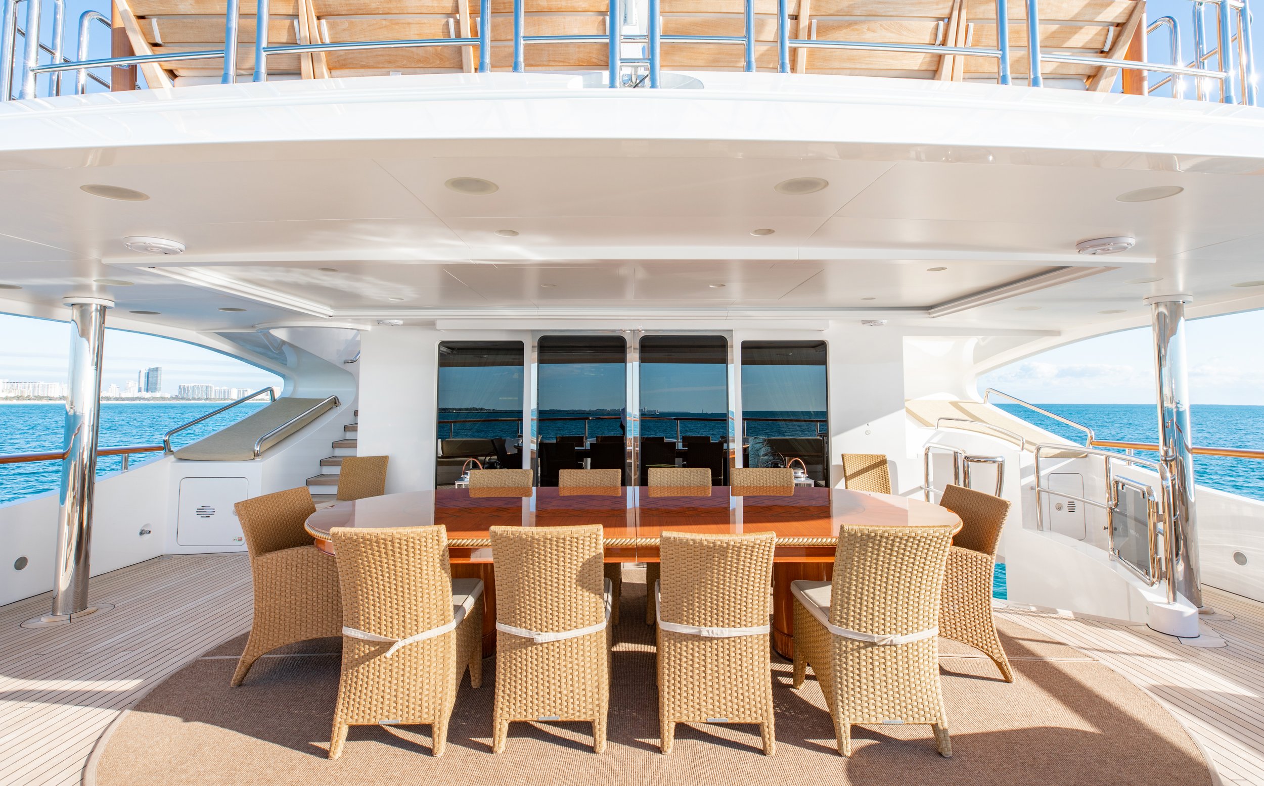 Outdoor dining area on a yacht with a large wooden table and wicker chairs, overlooking the ocean.