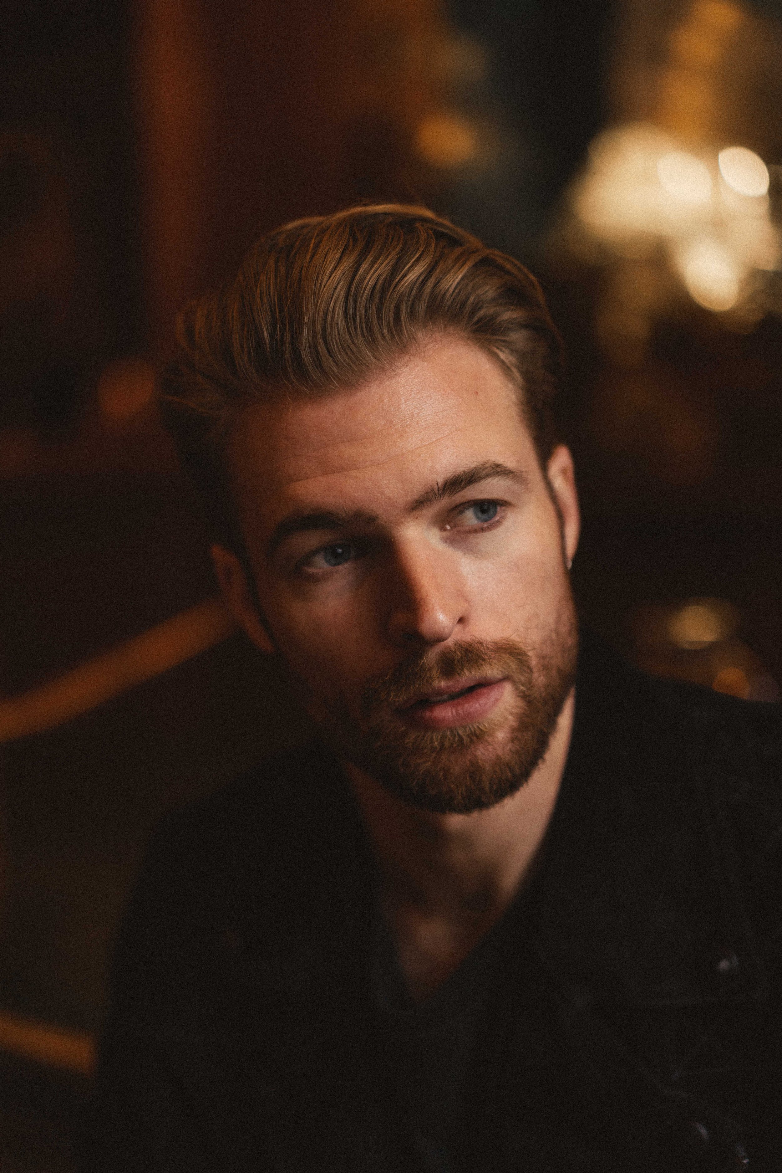 A close-up portrait of a young man with styled blonde hair and a beard, looking to the side, with dim warm lighting in the background.