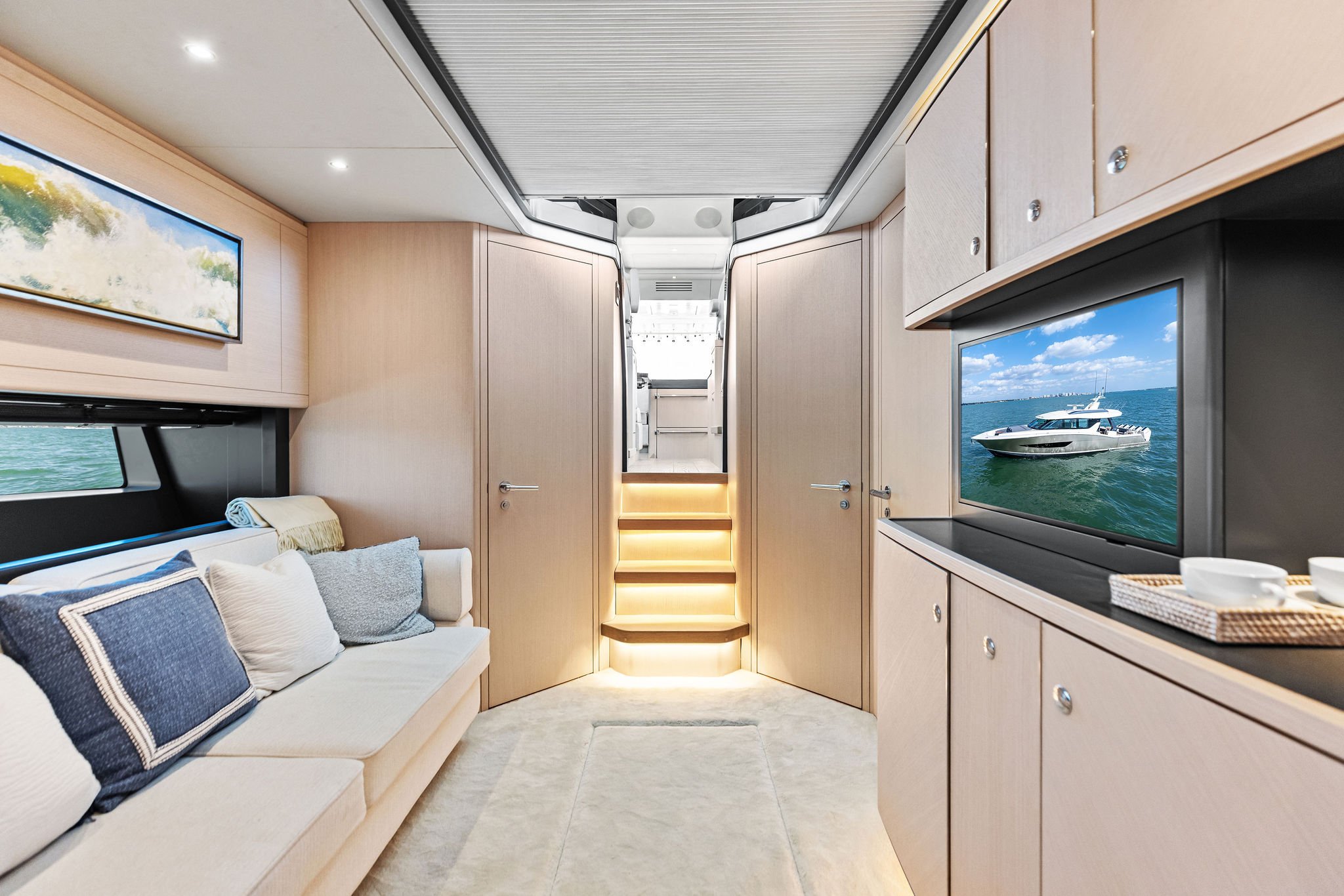 Interior of a luxury yacht with beige cabinetry, a cream sofa with blue and white pillows, a large flat-screen TV displaying a boat on water, and stairs leading to an upper deck illuminated with warm lighting.