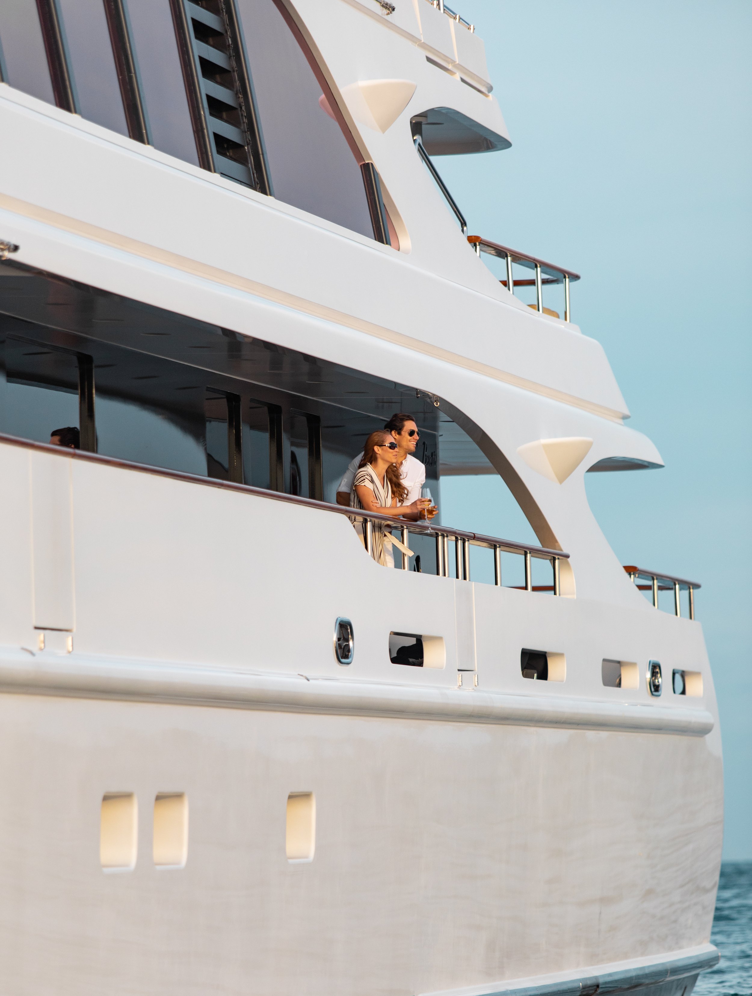 A large white yacht with two people on the deck, a man and a woman, both wearing sunglasses and enjoying drinks, against a clear blue sky.