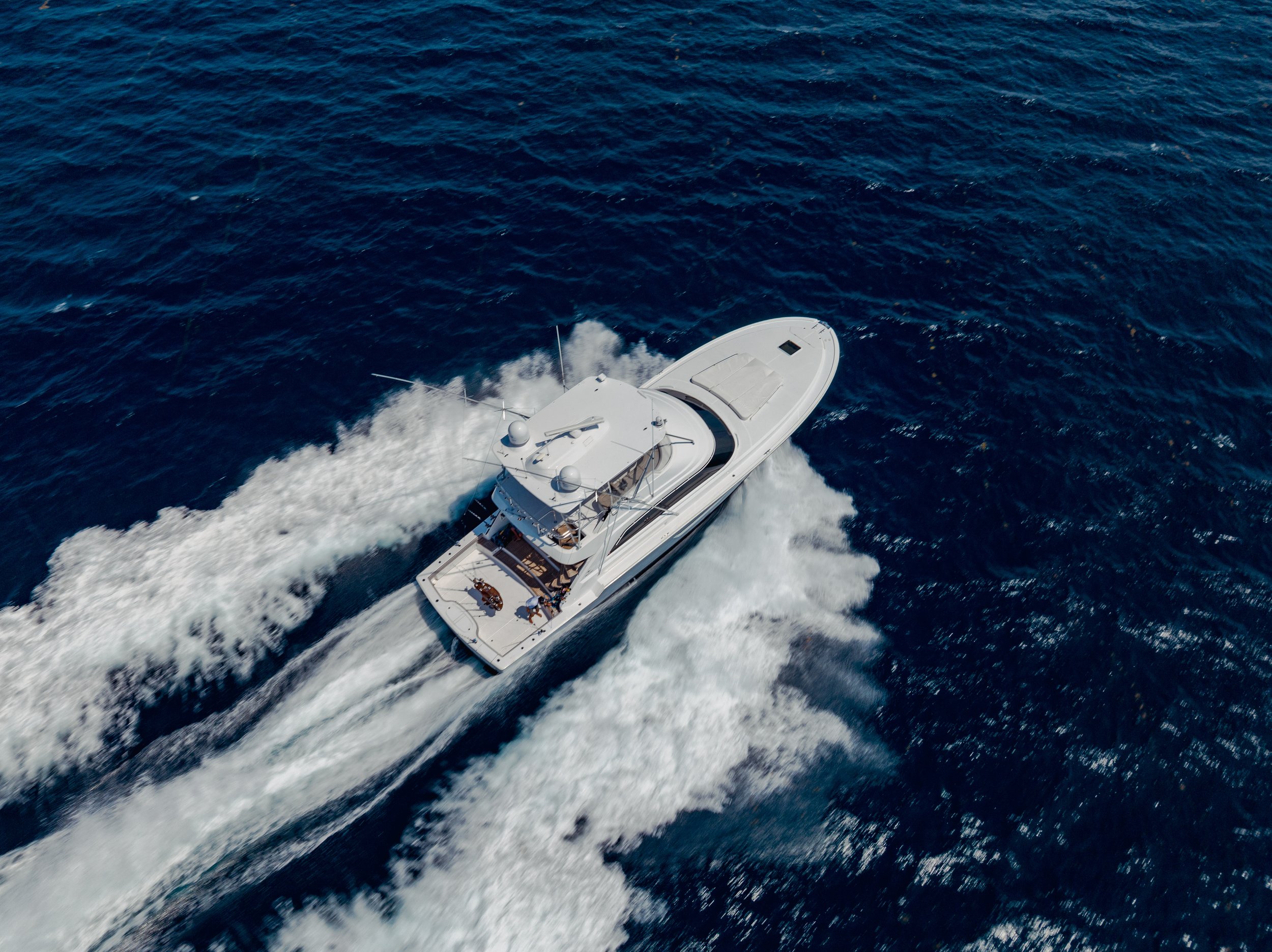 An aerial view of a white motor yacht speeding across deep blue ocean water, creating a foamy wake behind it.