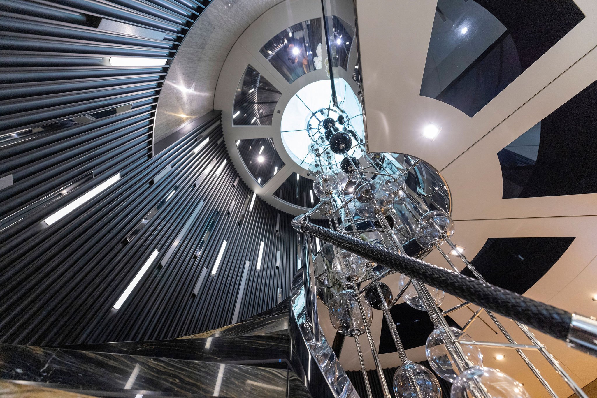 Interior view of a modern staircase taken from the bottom, looking up towards the ceiling, featuring a glass chandelier and black and white architectural elements.