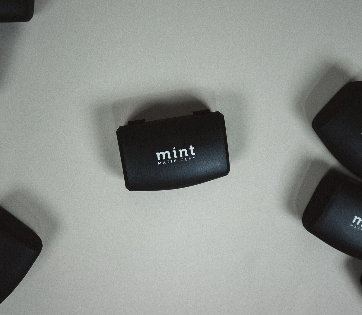 Black container labeled "mint" and "matte clay" on a light surface, surrounded by similar containers.