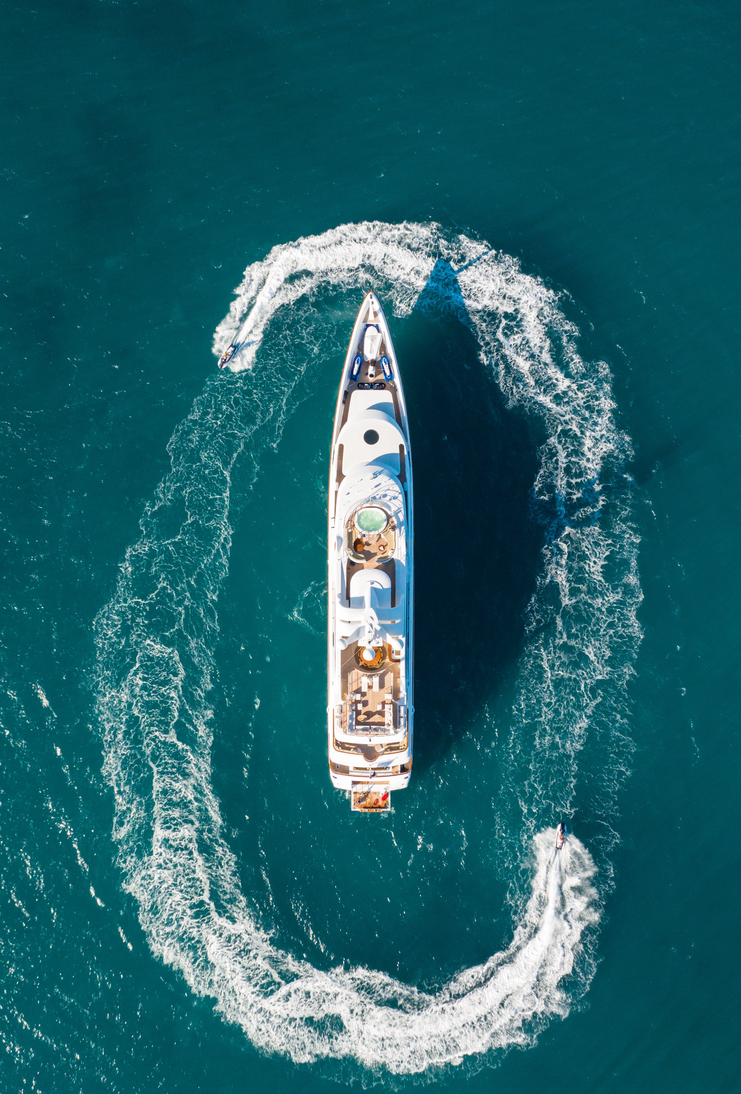 Aerial view of a large white yacht sailing in the water, with a smaller boat nearby, creating waves and trails in the ocean.