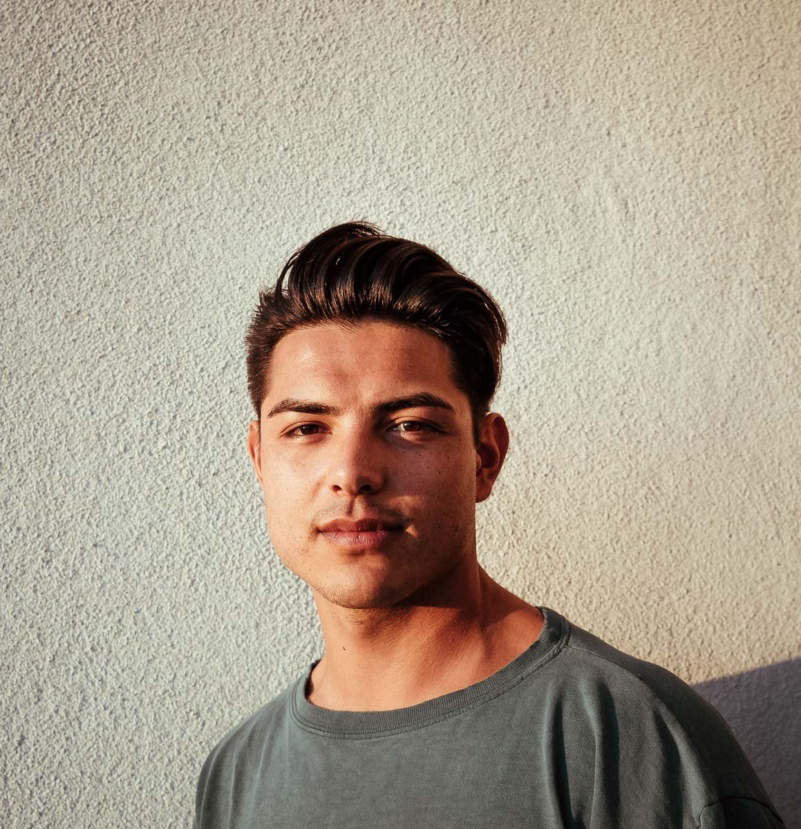 A young man with styled dark hair and light skin wearing a gray t-shirt, standing against a textured beige wall.