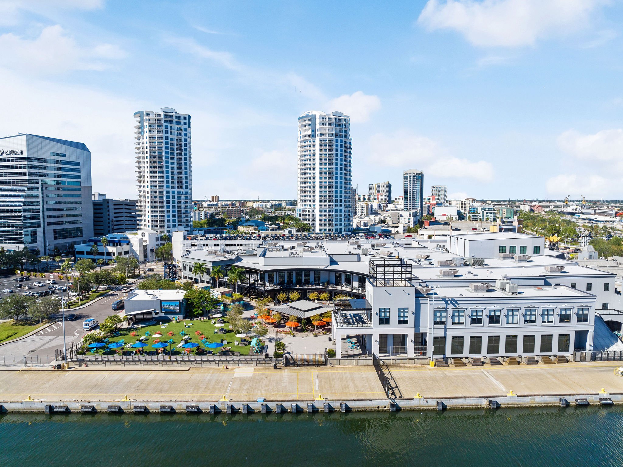 Aerial view of a modern urban area with high-rise buildings, commercial structures, a waterfront, and a recreational lawn with colorful umbrellas