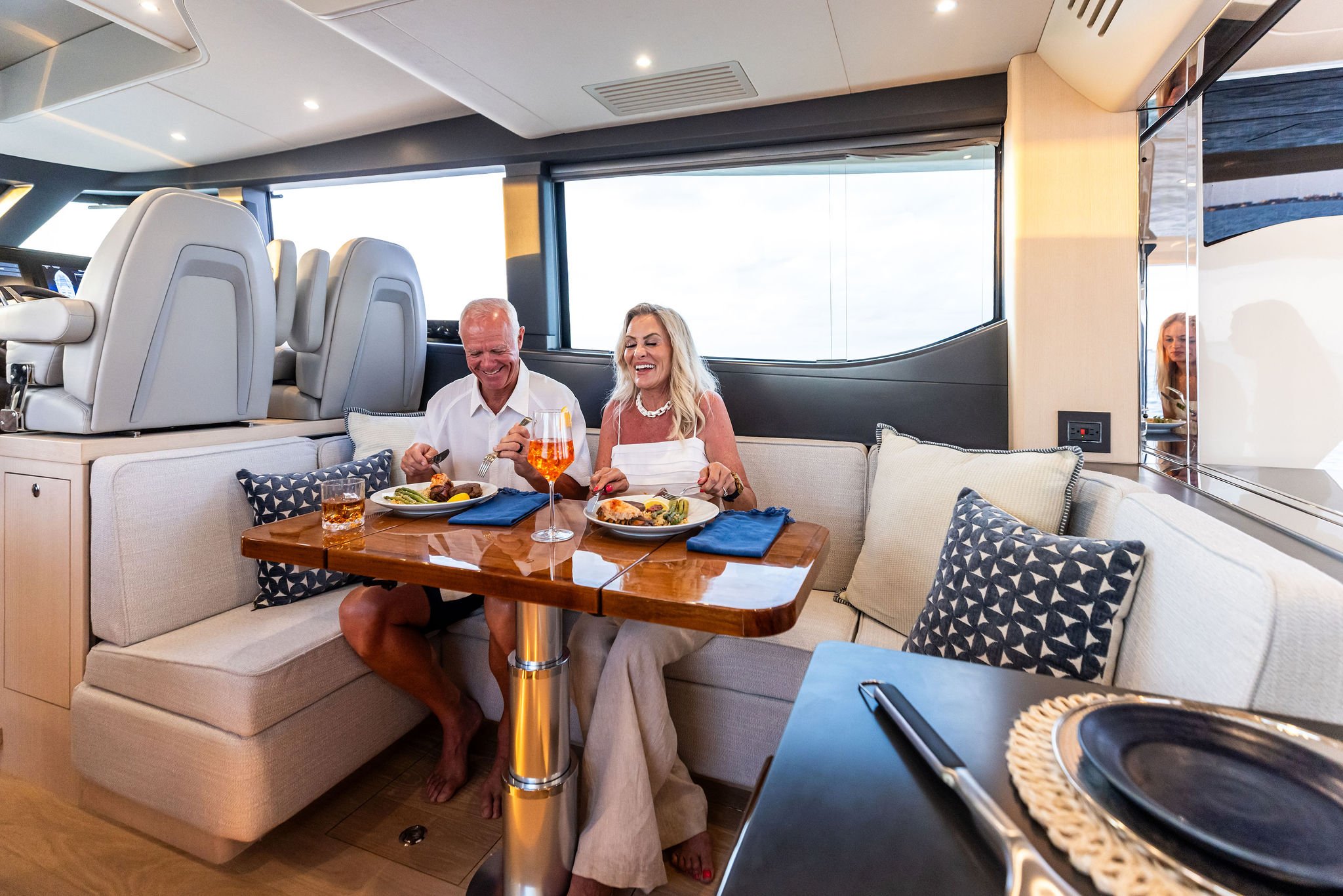 An older man and woman dining and laughing together on a yachting deck with large windows and a modern interior.